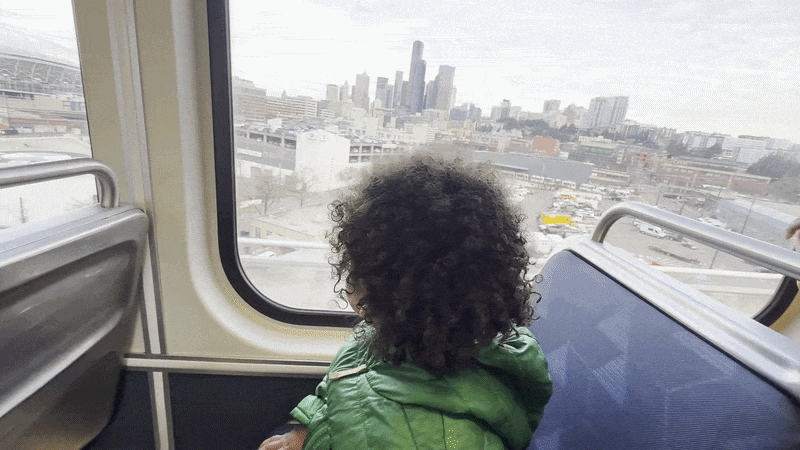 A curly haired small child sits on a blue seat on a train while pointing out the window at the Seattle skyline in the distance.