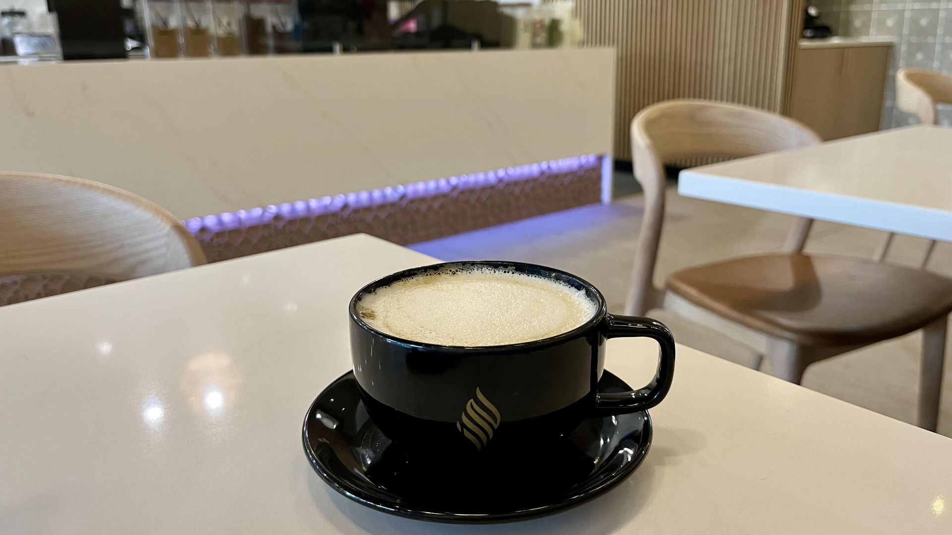 A black ceramic coffee cup with frothy latte on a matching saucer sits on a white table in a bright cafe, Haraz Coffee House in San Antonio. In the blurred background, there's a front counter with equipment and a wooden slat wall.