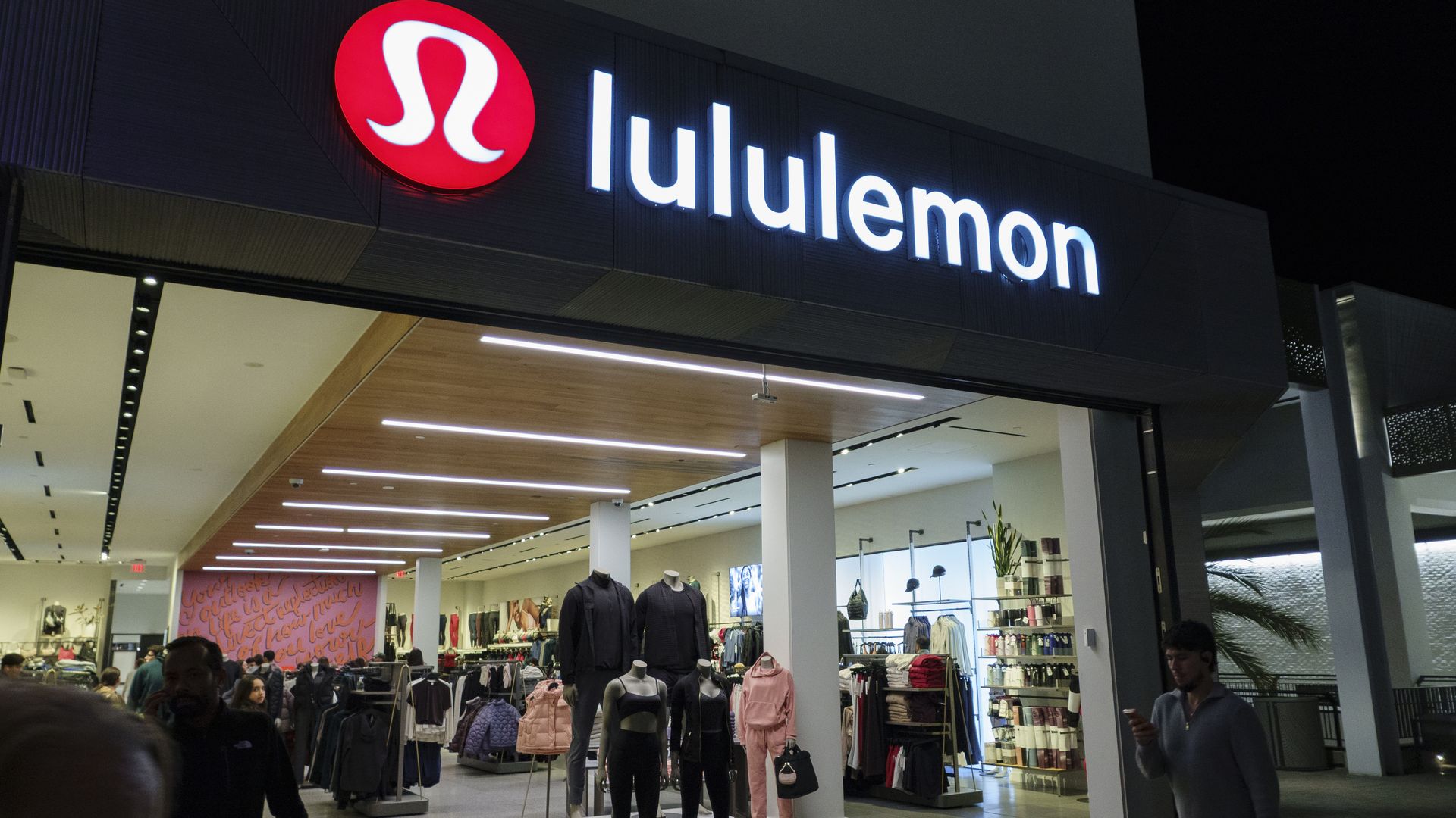 Night exterior of a lululemon store with a bright white lululemon sign and red circular logo; interior shows mannequins and clothing racks with shoppers.