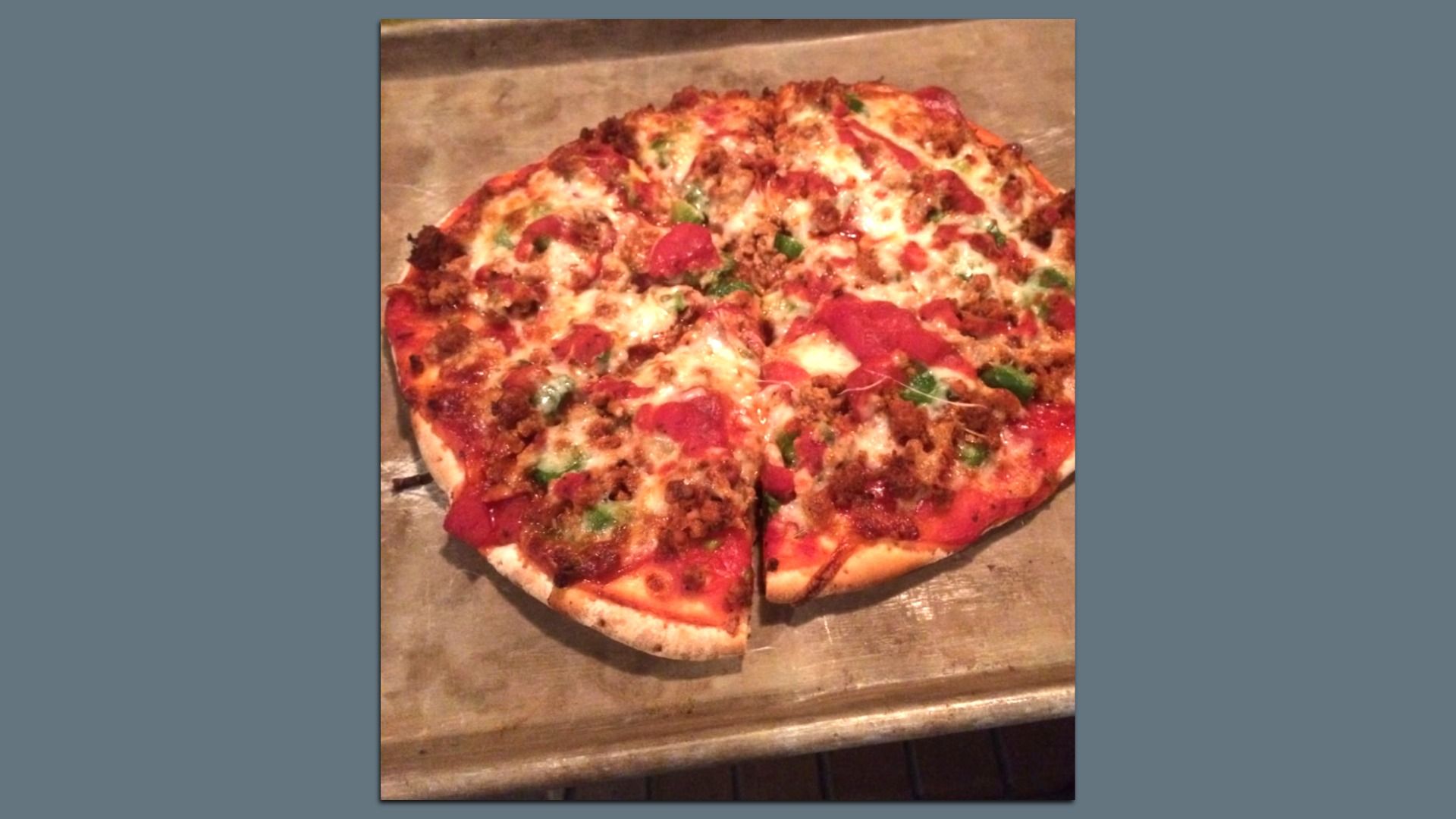 A large pizza on a baking sheet with melted cheese, pepperoni, sausage crumbles, and chopped green peppers on a thick crust.