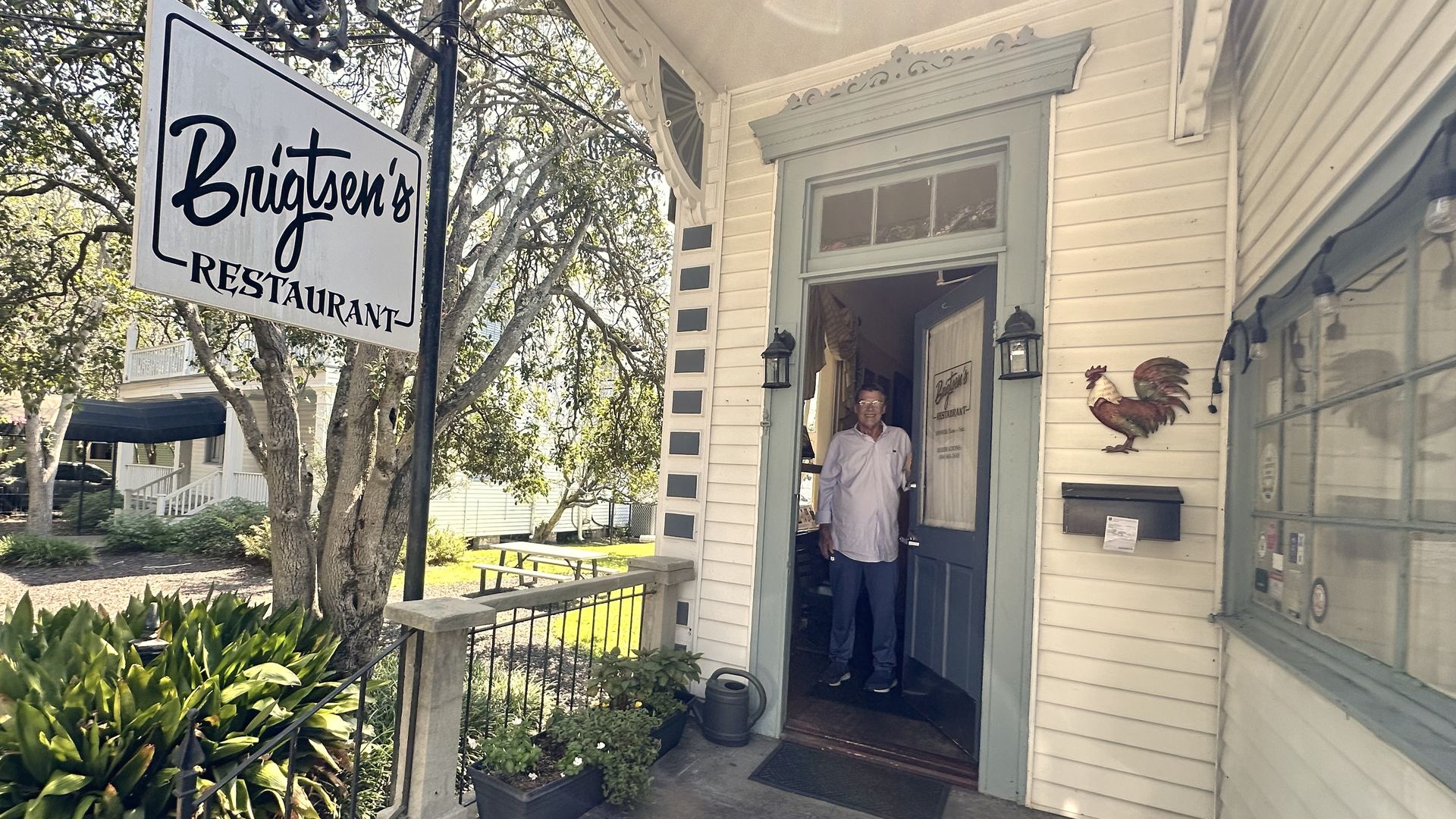 Street view of a small restaurant with a pale blue doorway reading "Brigtsen's". An older man stands in the open door. A large sign on a pole says "Brigtsen's Restaurant". A rooster mural and lush greenery surround the entrance.