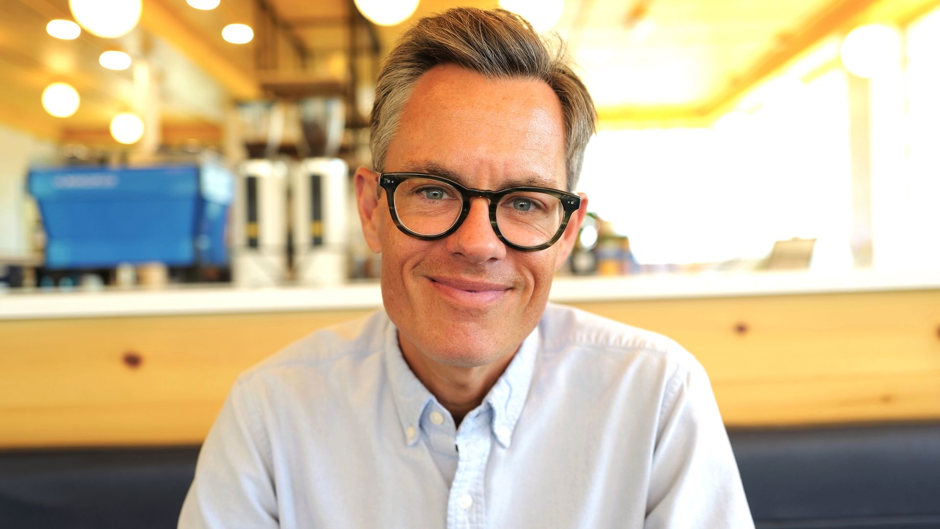 A photo of a man in glasses sitting in a bright coffeehouse. 