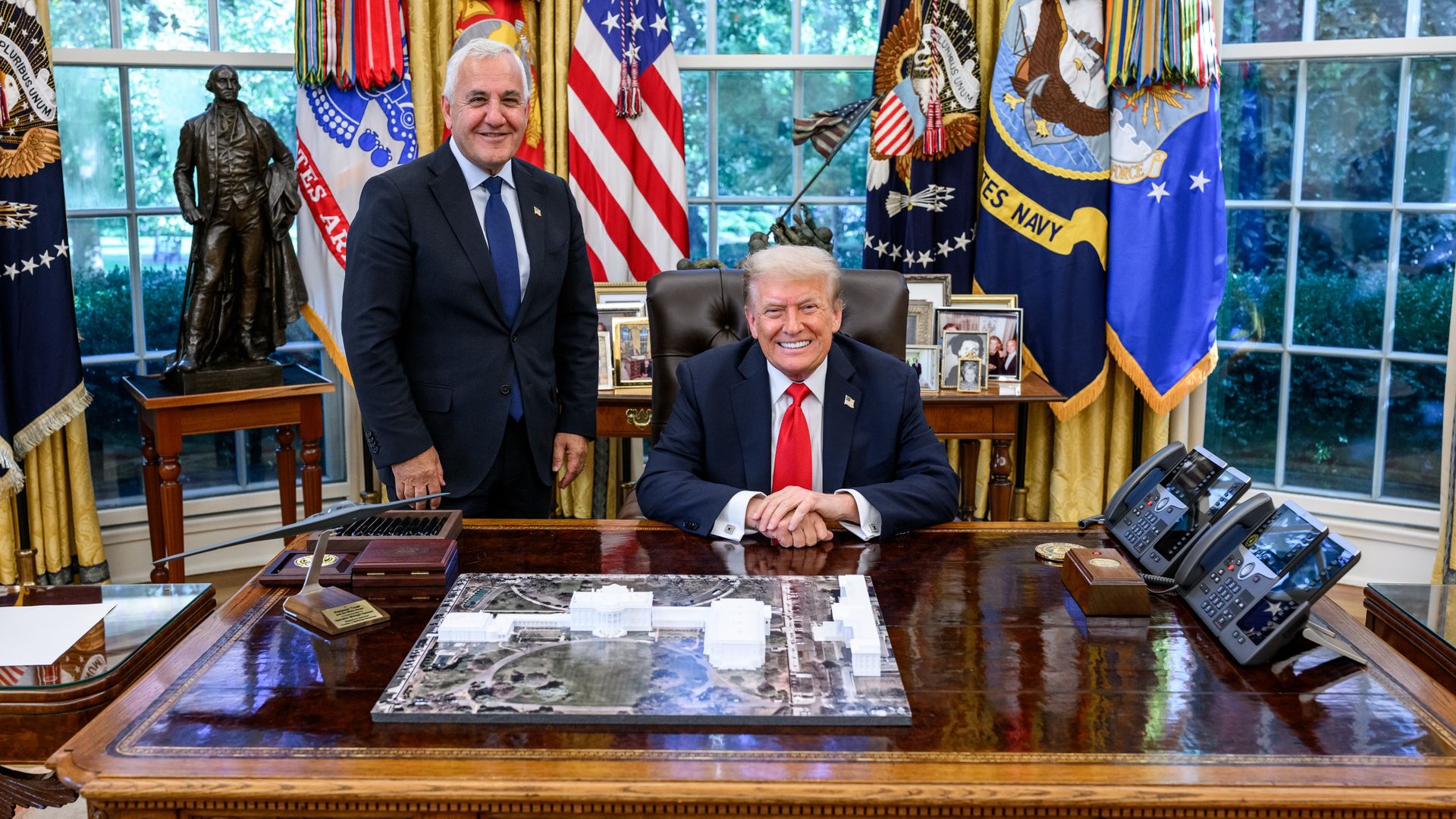 Two men in suits in an official office with American flags behind them; one seated with red tie and the other standing with blue tie near a wooden desk with phones, model, and photo frames.