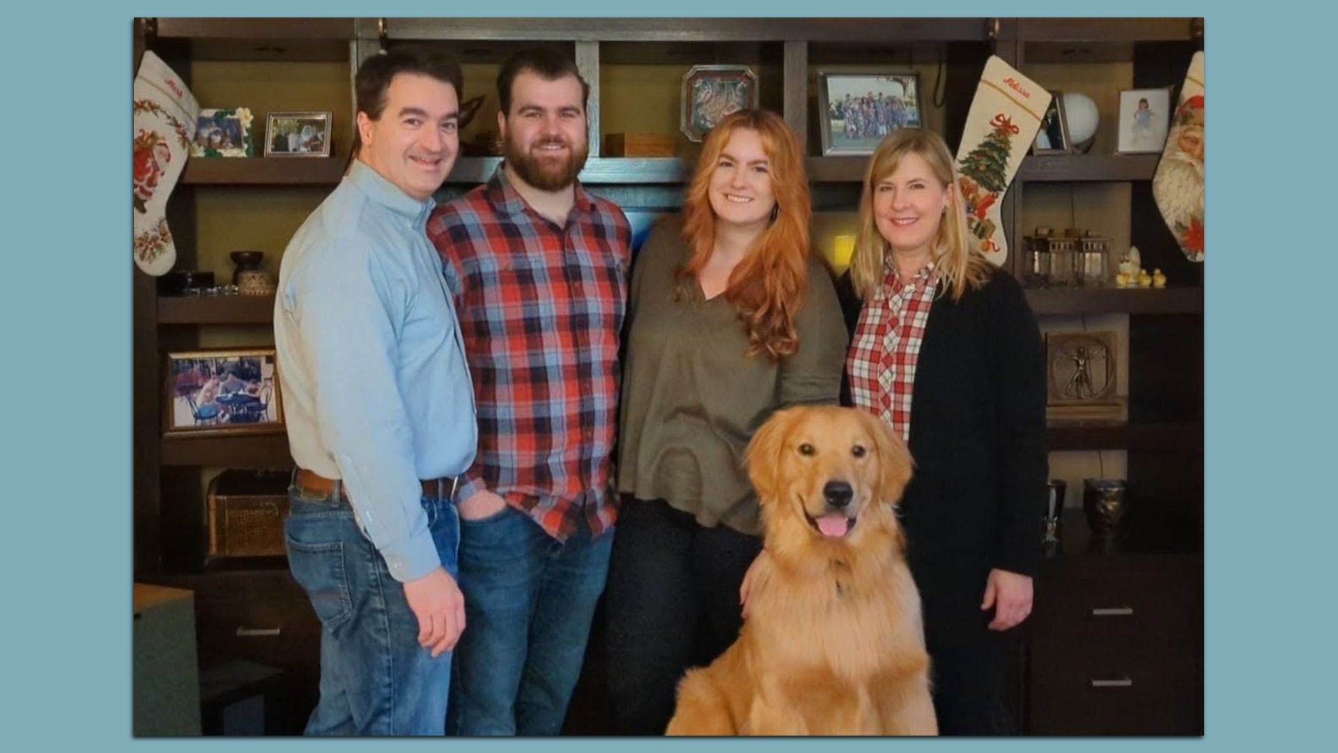 Four people (two men, two women) pose with a golden retriever in a home living room; festive stockings, shelves with photos frame the cozy scene.
