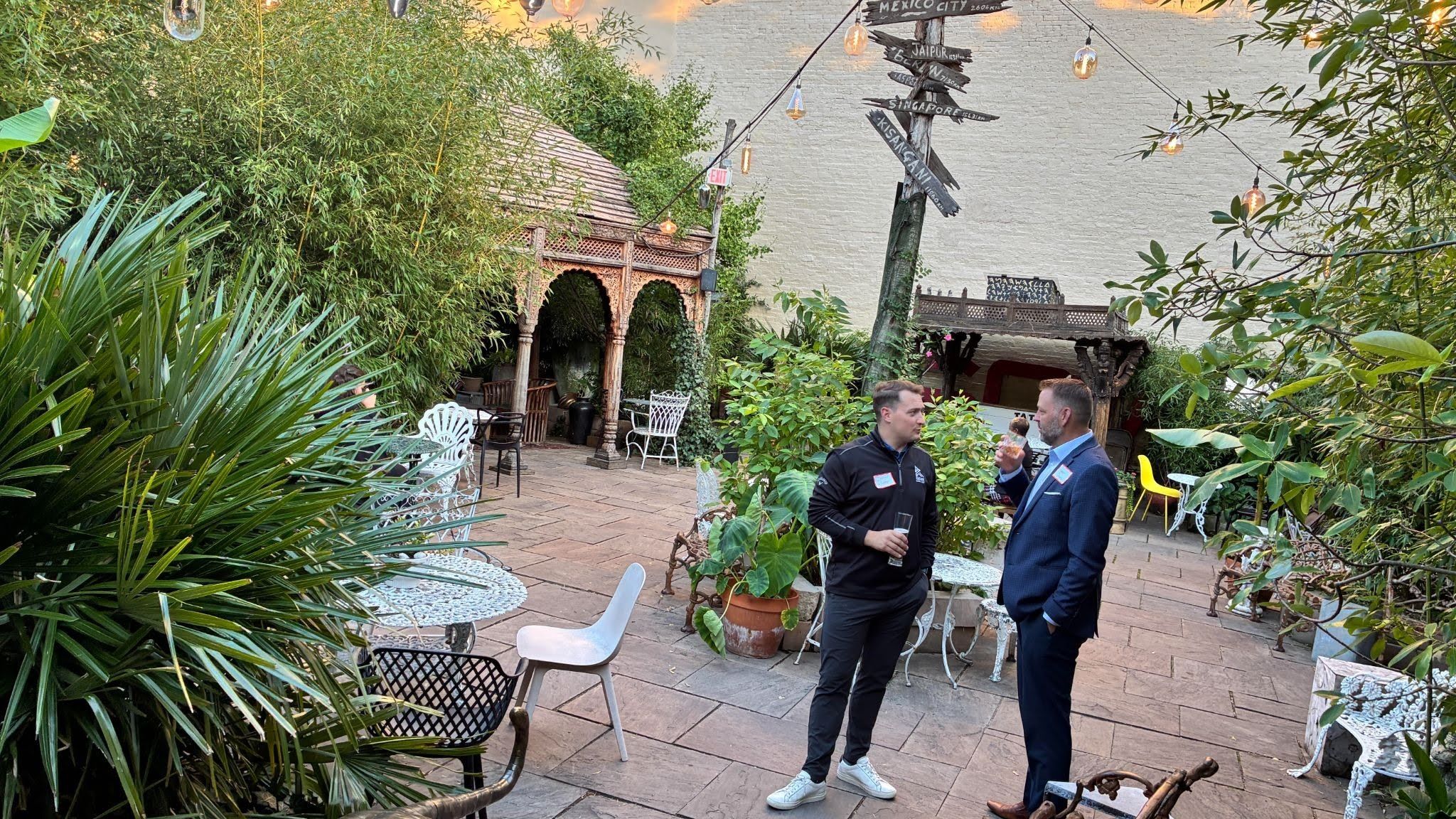 Lush garden courtyard with tall bamboo, potted plants, and ornate wooden gazebo. String lights hang overhead as two men in smart attire chat, one holding a glass, near metal chairs and a signpost.