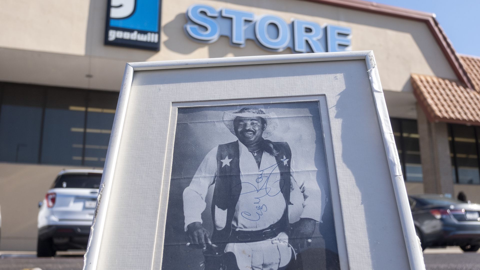 Black-and-white framed photo of a smiling man in a cowboy outfit with star badges, propped up outside a Goodwill store under a bright blue sky.