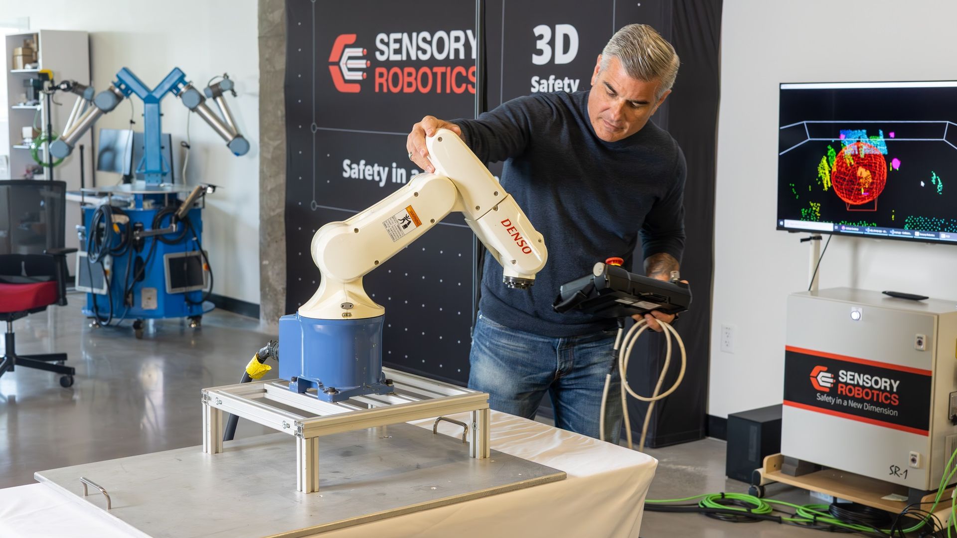 A man in a dark shirt adjusts a white robotic arm on a metal lab table; behind him is a banner that reads "SENSORY ROBOTICS" and "3D Safety," while a monitor shows a colorful 3D display.