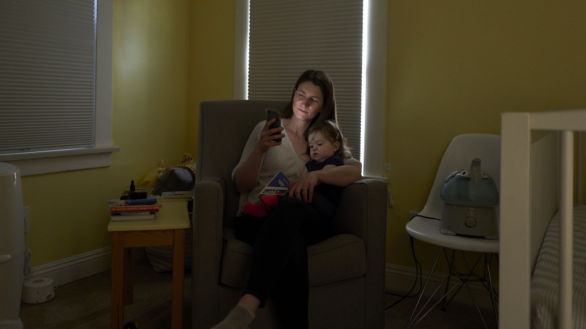 In this image, a mother looks at her phone while holding her infant in her lap in a chair in the corner of a nursery.