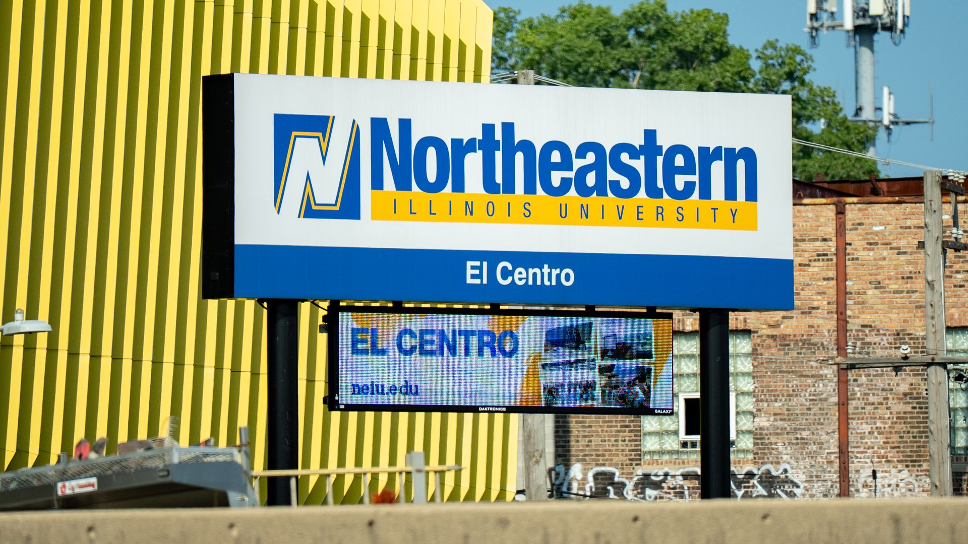 Northeastern Illinois University billboard with "El Centro" in white type on a blue background. The sign is in front of a bright yellow building.