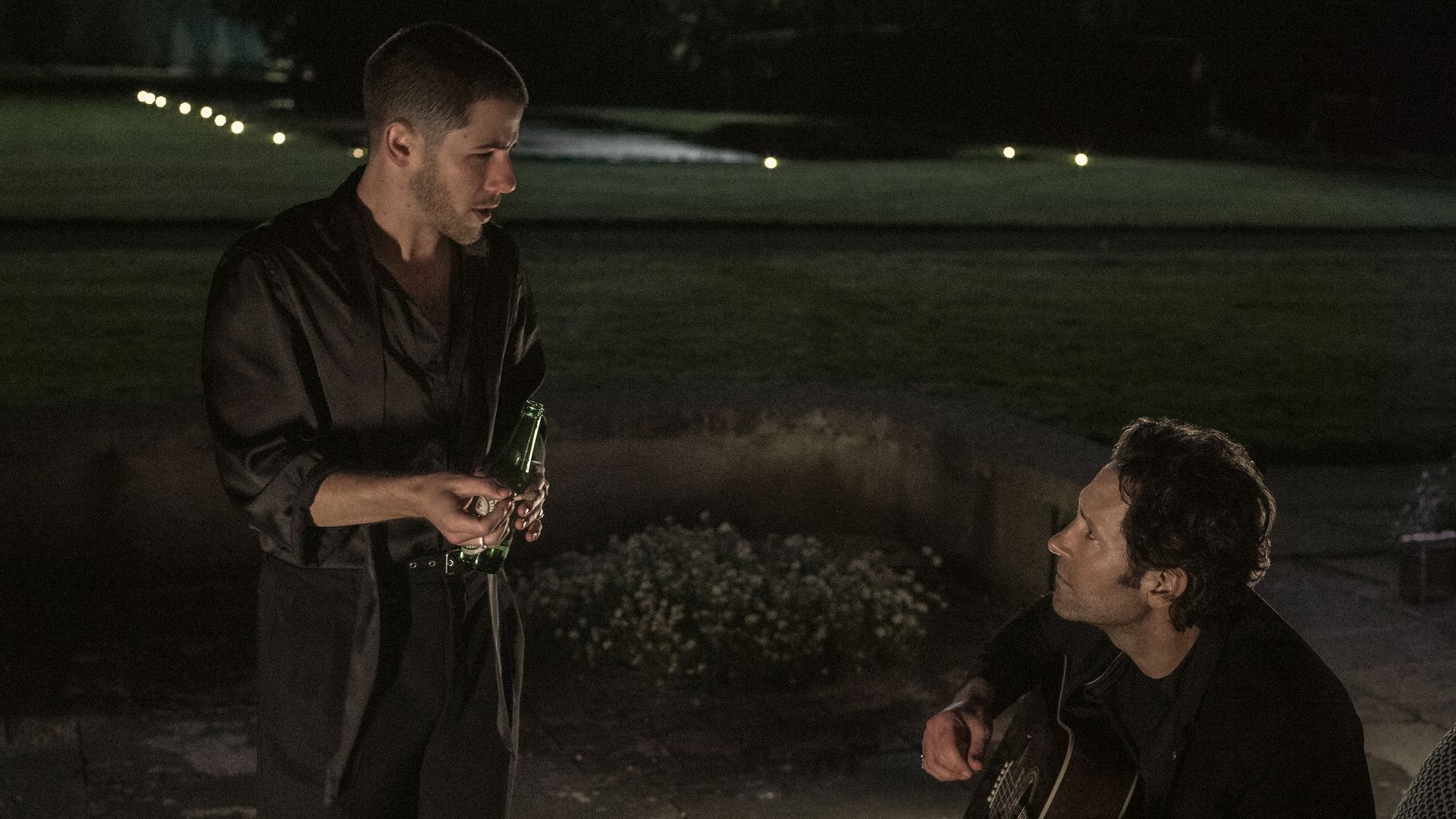 Two men at night outdoors; one stands holding a green bottle in black clothing, the other sits with an acoustic guitar and looks up at him, as if singing, with soft garden lights in the distance.