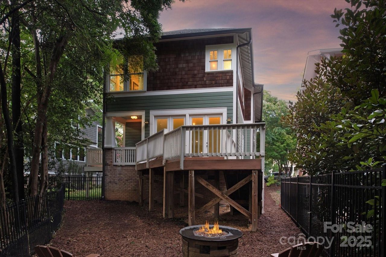 Two-story house with green and brown siding, white-trimmed windows lit warmly inside, elevated wooden deck, fenced yard with trees, and fire pit at dusk.