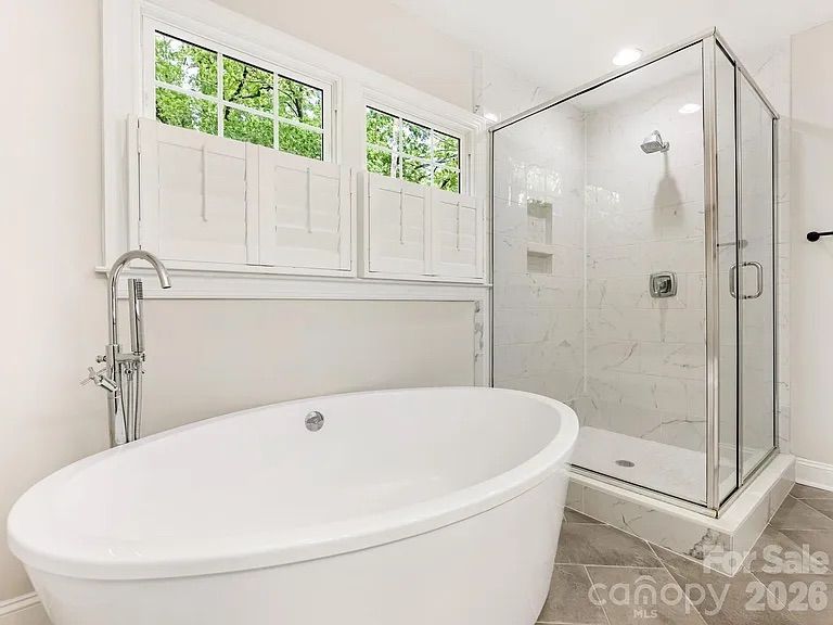Bright modern bathroom with a white freestanding oval tub, chrome floor faucet, a glass-enclosed marble shower, and a large window with white shutters overlooking green trees.