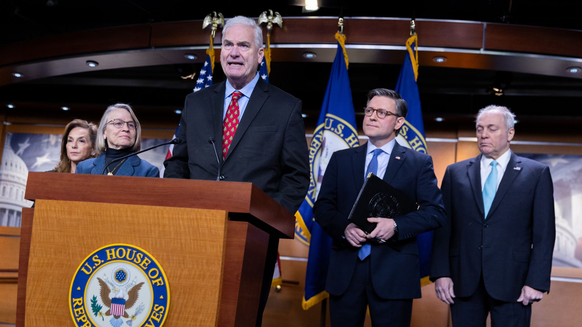 Five people in suits at a U.S. House of Representatives podium with flags behind them in a formal setting, one man speaking while others listen attentively.