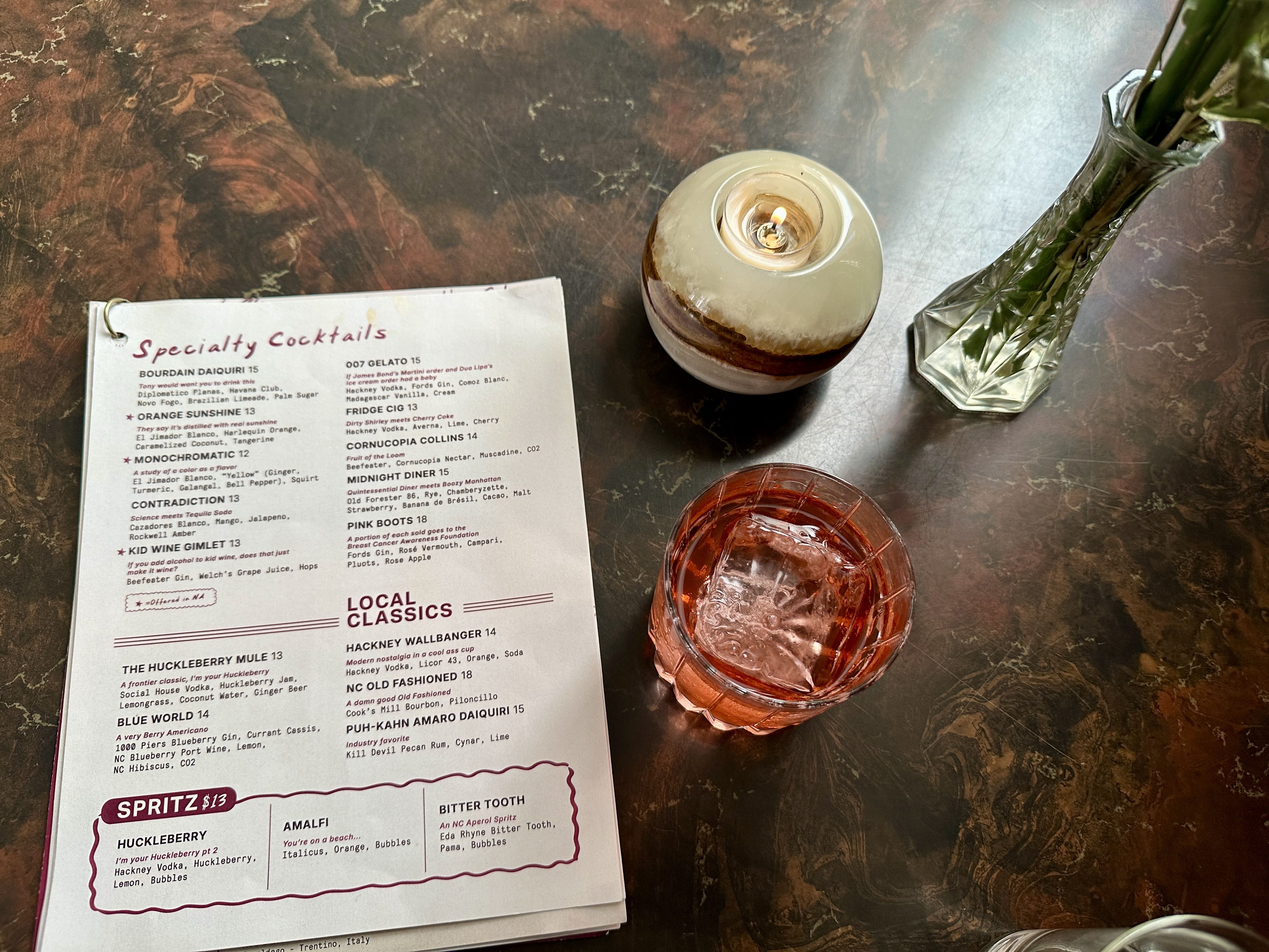 Top-down view of a marble table: a lit ceramic candle holder in the center, a menu titled "Specialty Cocktails" on the left, and a faceted glass vase with green stems on the right; pink cocktails nearby. Huckleberry NoDa now open.
