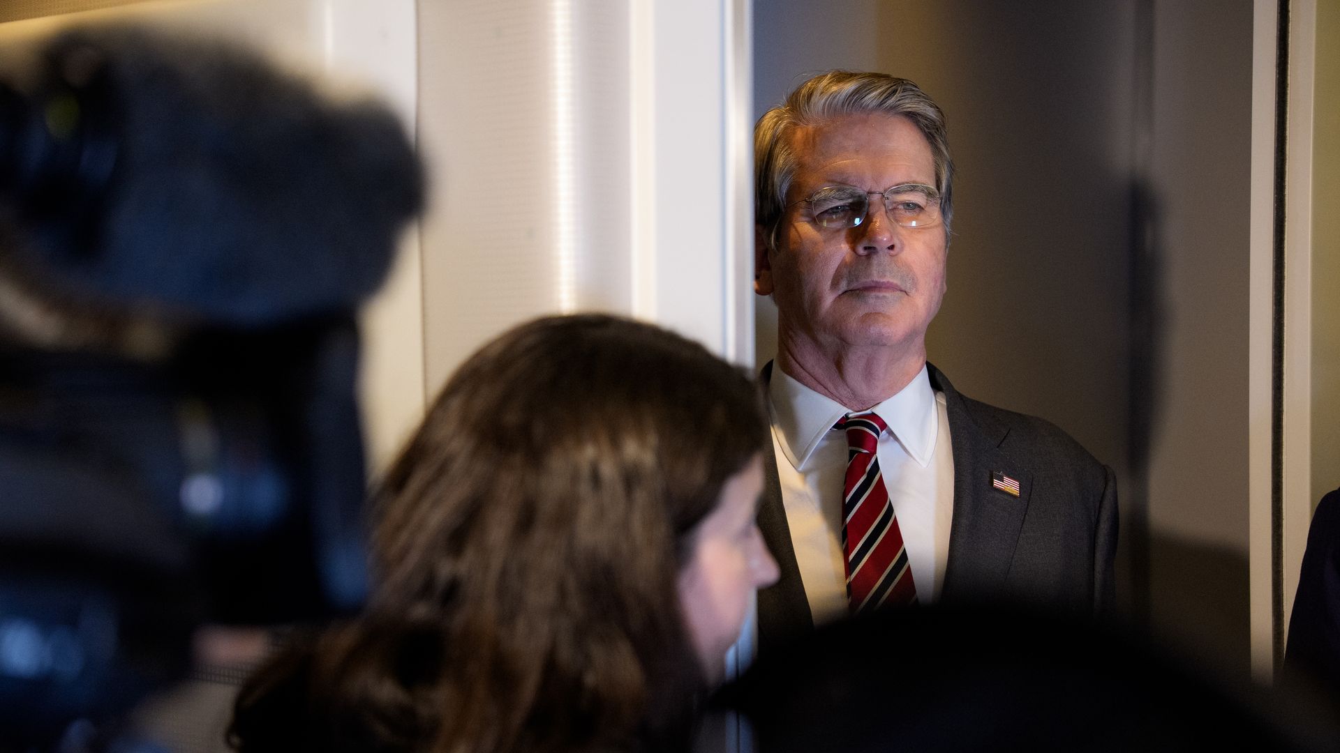 Scott Bessent — wearing glasses, a striped red tie, a dark suit, a collared shirt and an American flag pin — stands in a doorway.