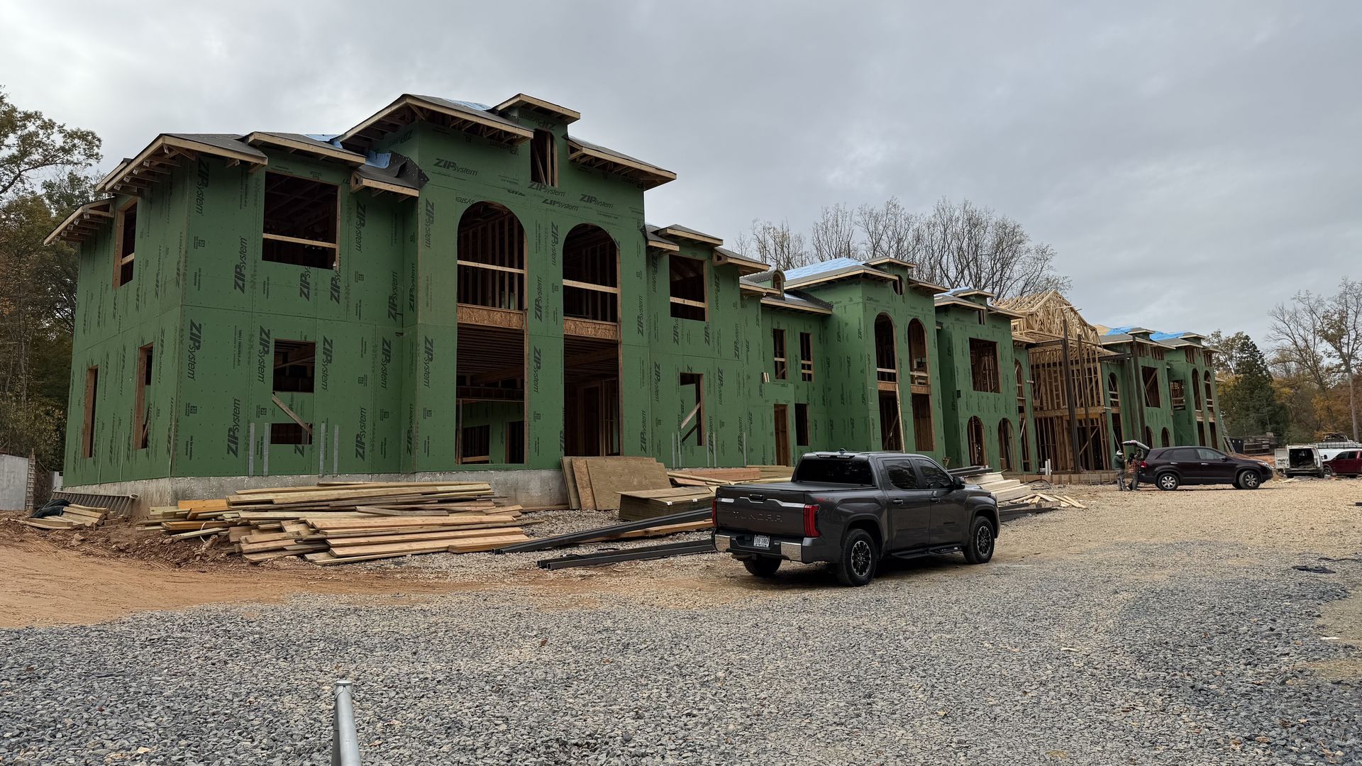 Large two-story building under construction with green ZIP System sheathing, exposed wooden framing, several trucks parked, gravel and wood materials on site, cloudy sky overhead.