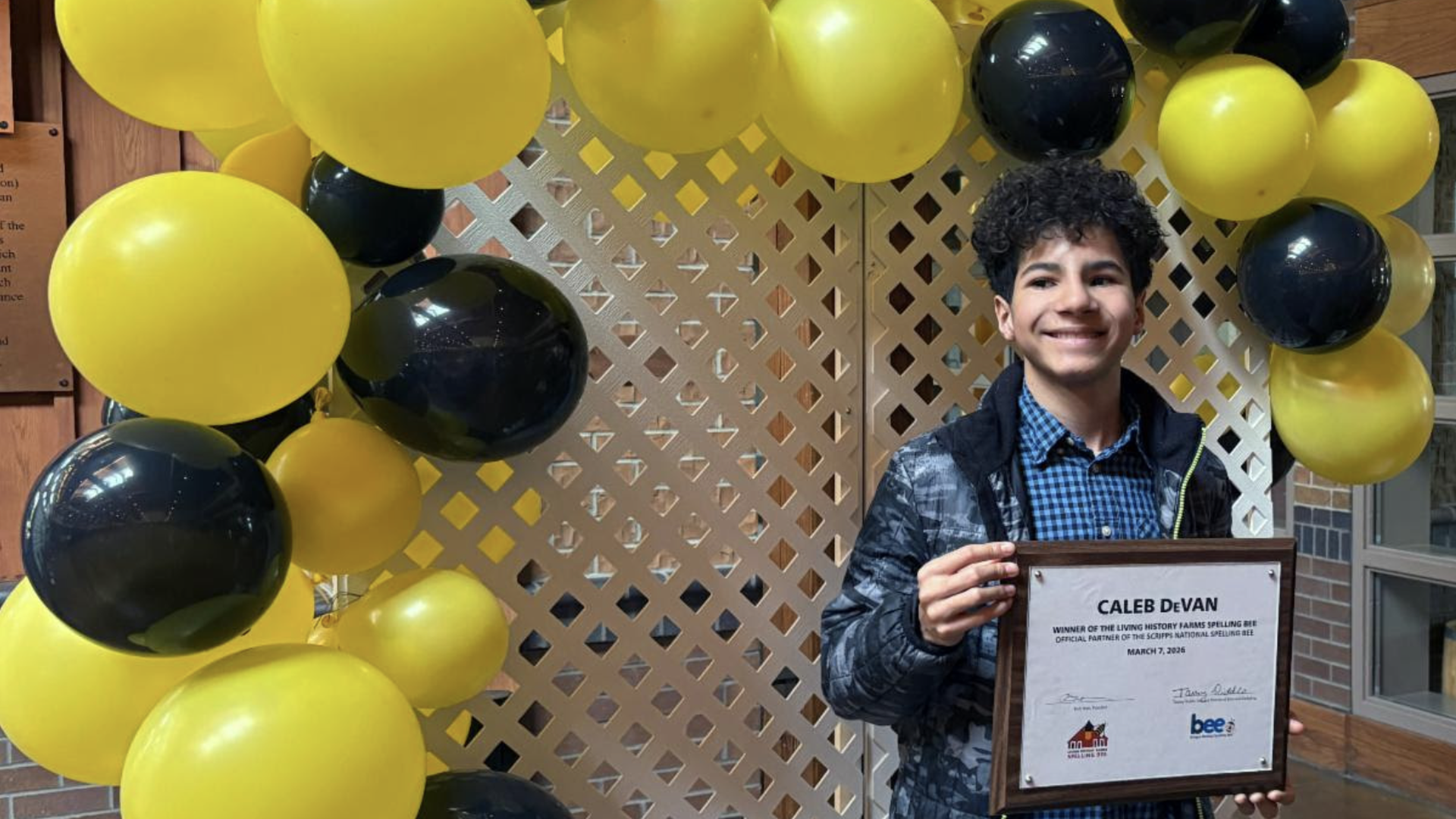 Smiling young man stands beside a yellow and navy balloon arch, holding a framed certificate. He wears a blue checkered shirt and dark jacket, in front of a lattice backdrop.