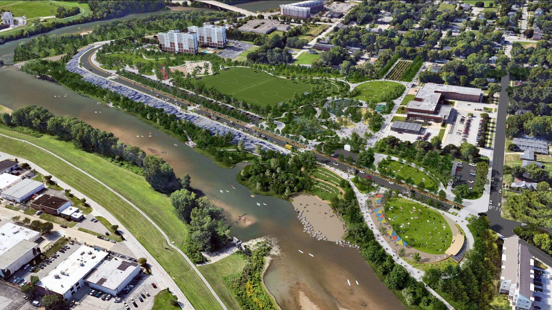 An aerial view of a riverfront park: a green soccer field with surrounding paths and trees, a curved road along the river, nearby apartments, a beach area, and colorful tents at the bottom.