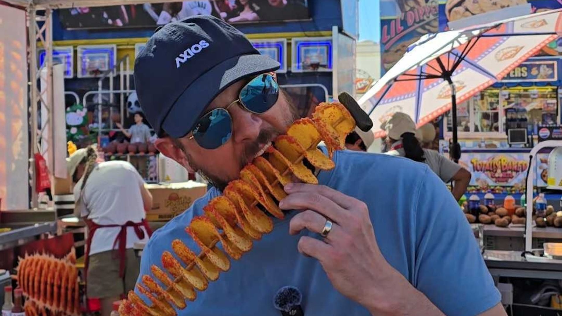 Man in a blue T-shirt and cap with mirrored sunglasses bites into a long spiral-cut fried potato on a stick at a colorful carnival food stand.