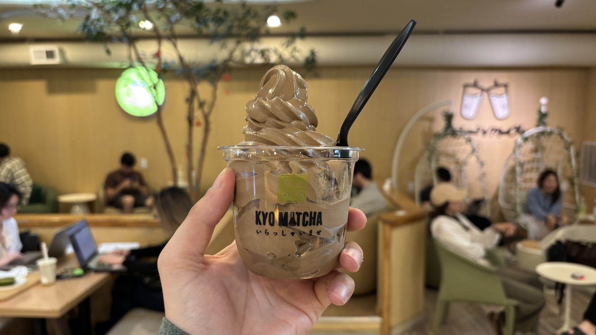 Hand holding a cup of chocolate soft serve ice cream with a black spoon inside, branded with "KYO MATCHA" in a cozy cafe with people sitting and chatting in the background.