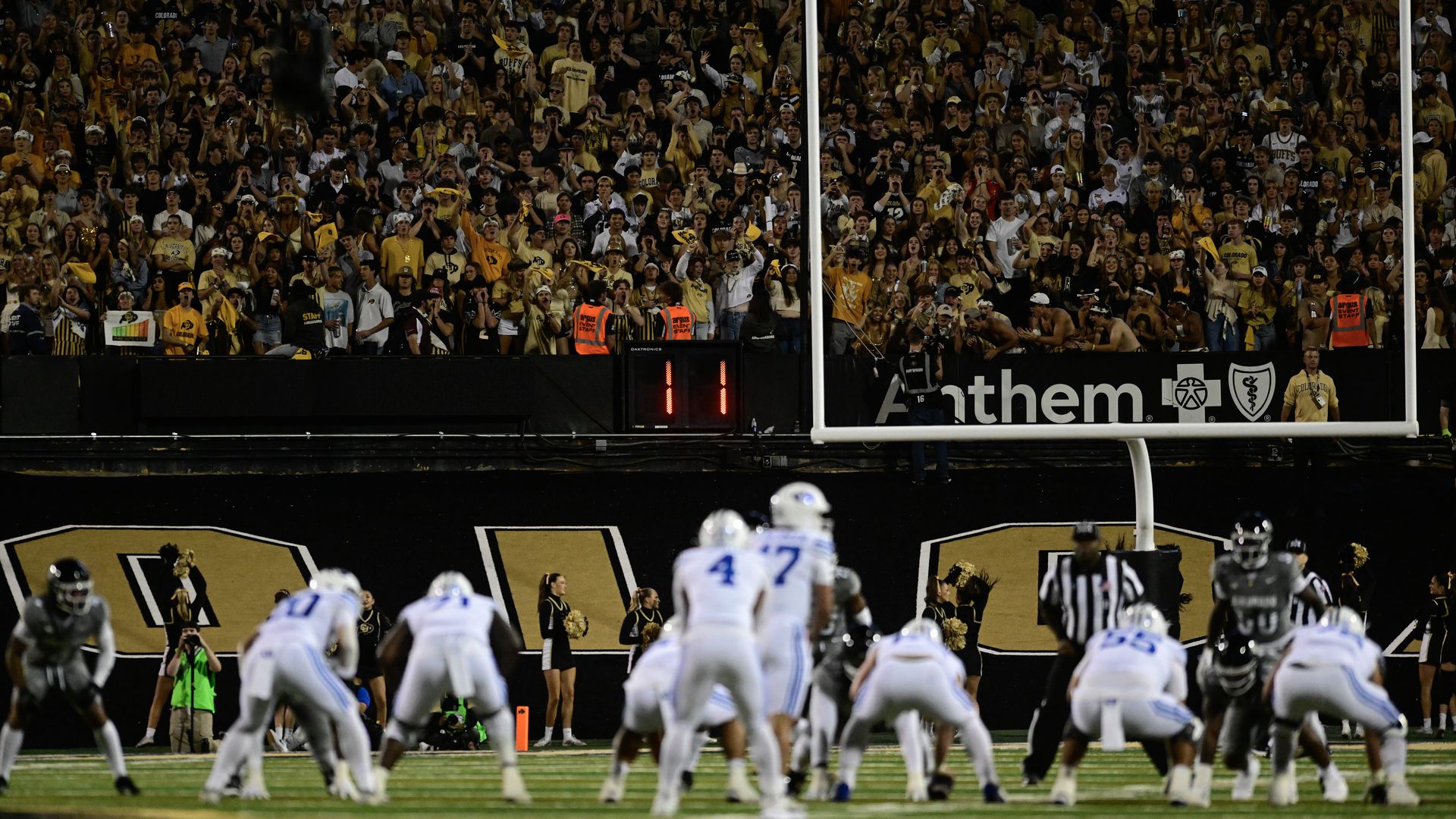 CU versus BYU football game with players in white and gray uniforms on the field, large crowd in yellow and black in stands.
