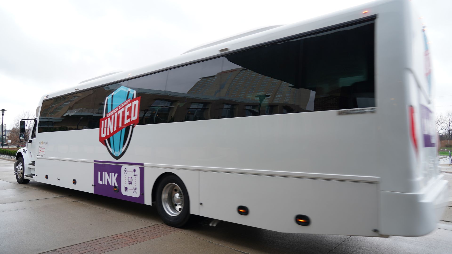 White tour bus with dark tinted windows reflecting buildings, featuring a large red United shield logo and a purple Link panel along the lower side.