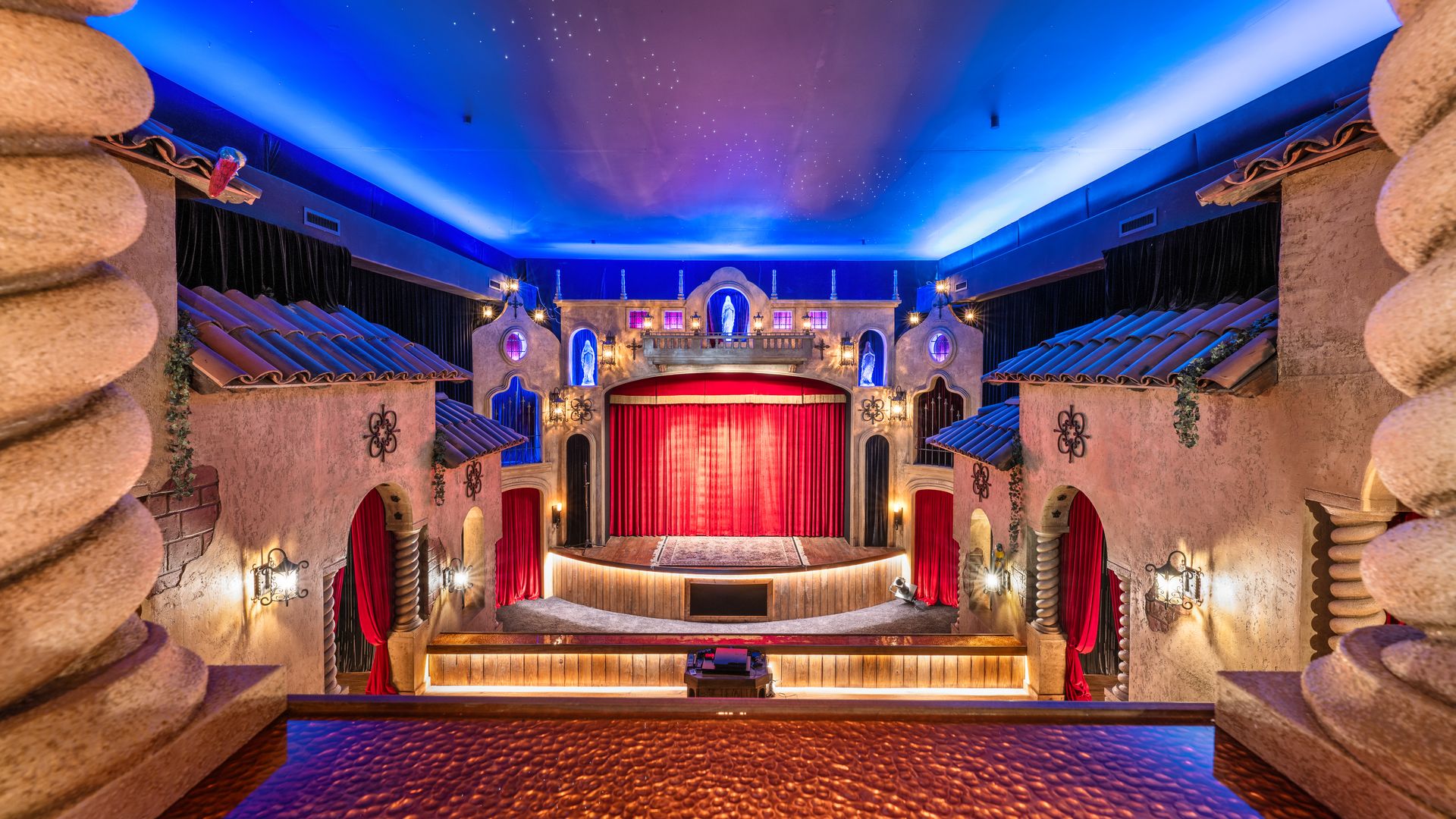 Interior of a theater with red curtains on stage, blue ambient lighting on ceiling, rustic stucco walls, and ornamental columns framing the view.
