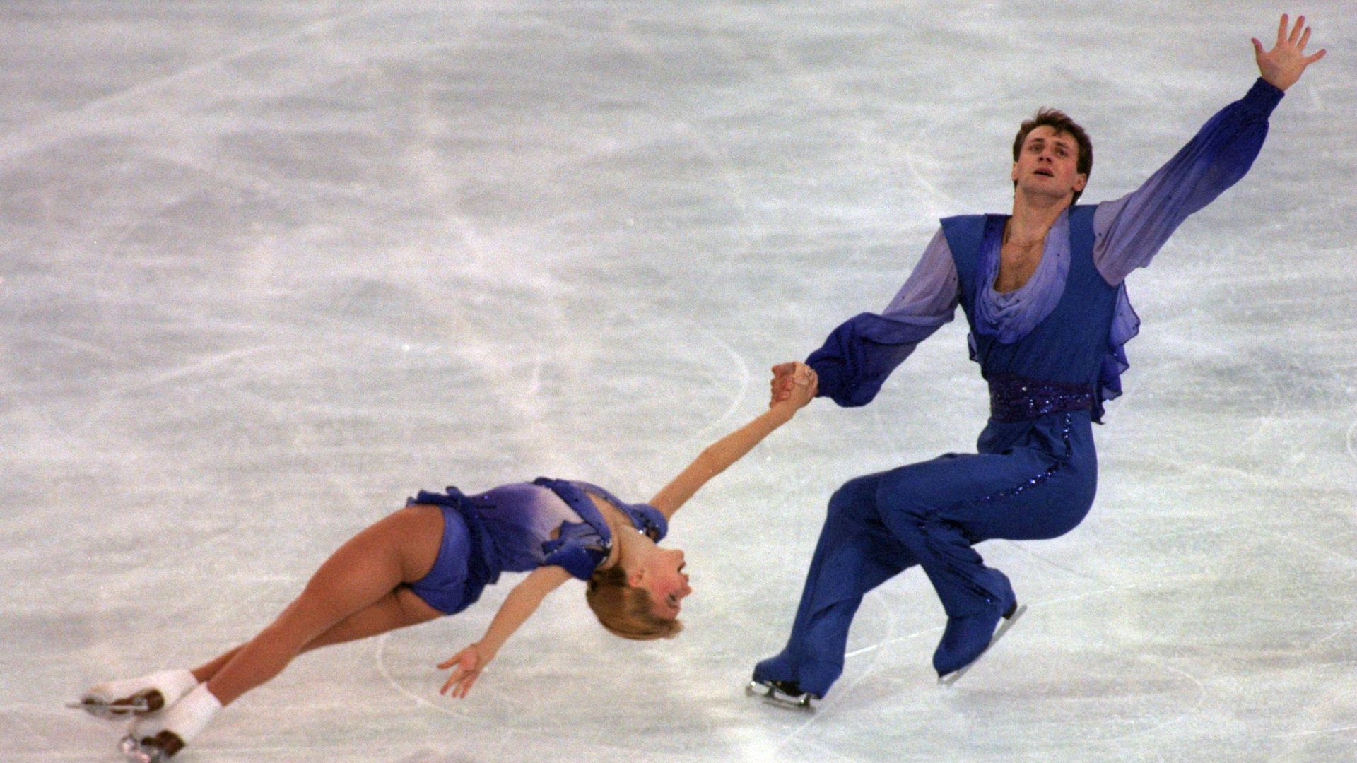 A photo showing two ice skaters wearing purple, sparkly outfits. The man to the left has one hand reaching up and the other grasping a woman who is bending back, skating with. her legs crossed.