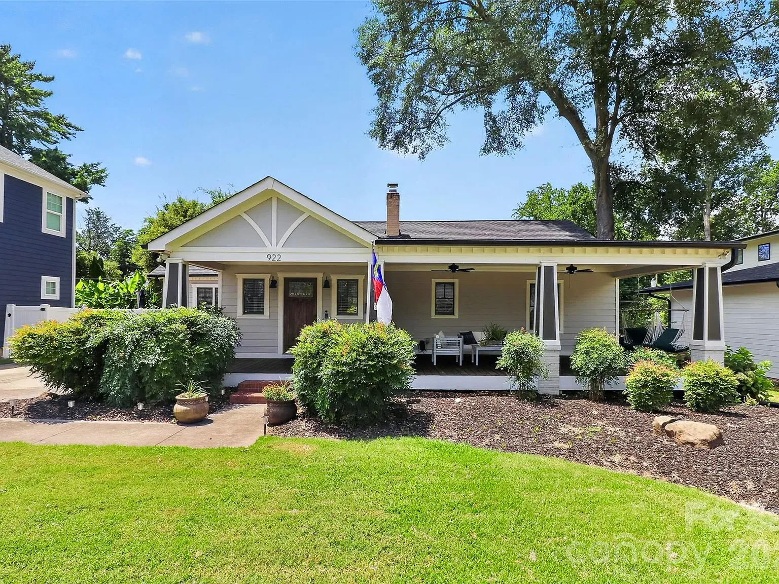 One-story ranch-style house with light gray siding and white trim, a covered front porch with pillars, and a flag by the door. Front yard has shrubs, mulch beds, and a large tree under a blue sky.