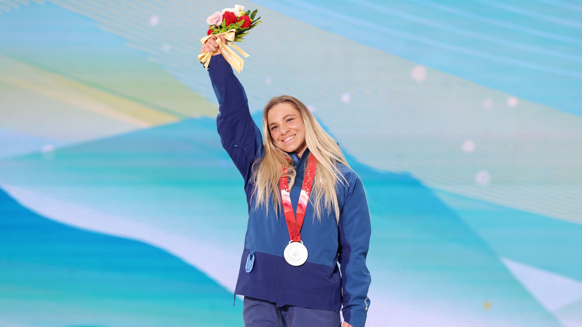 Smiling female athlete with long blonde hair wearing a blue USA jacket and pants, holds a silver medal and raises a bouquet of flowers against a blue-green abstract background.