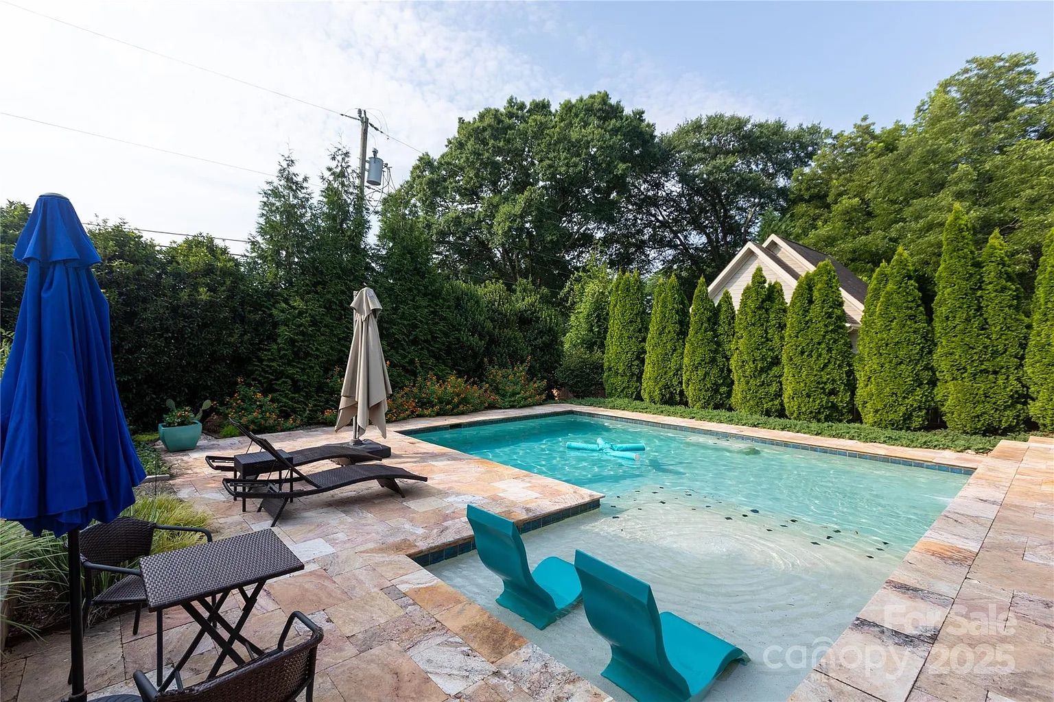 Outdoor pool surrounded by tile deck, with two turquoise lounge chairs submerged in shallow water, two closed umbrellas (blue and beige), lounge chairs, and lush green trees and shrubs around.