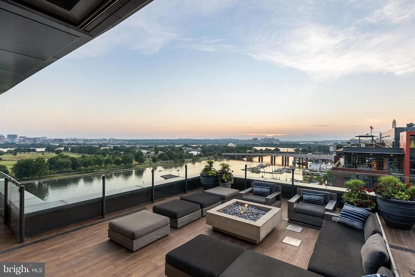 Rooftop terrace with gray cushioned sectional seating around a square fire pit, glass railing, and large planters. A river, park, and distant city skyline glow at sunset.