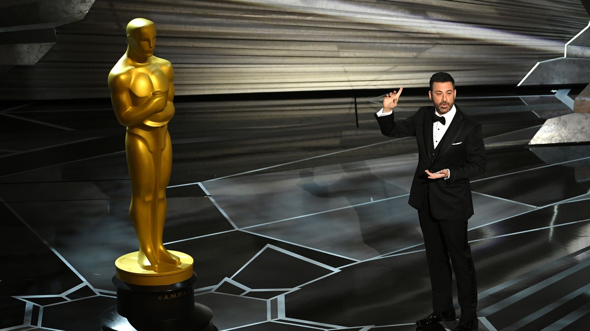 Jimmy Kimmel onstage at the Academy Awards, with a finger raised, speaking to a giants Oscar statue.