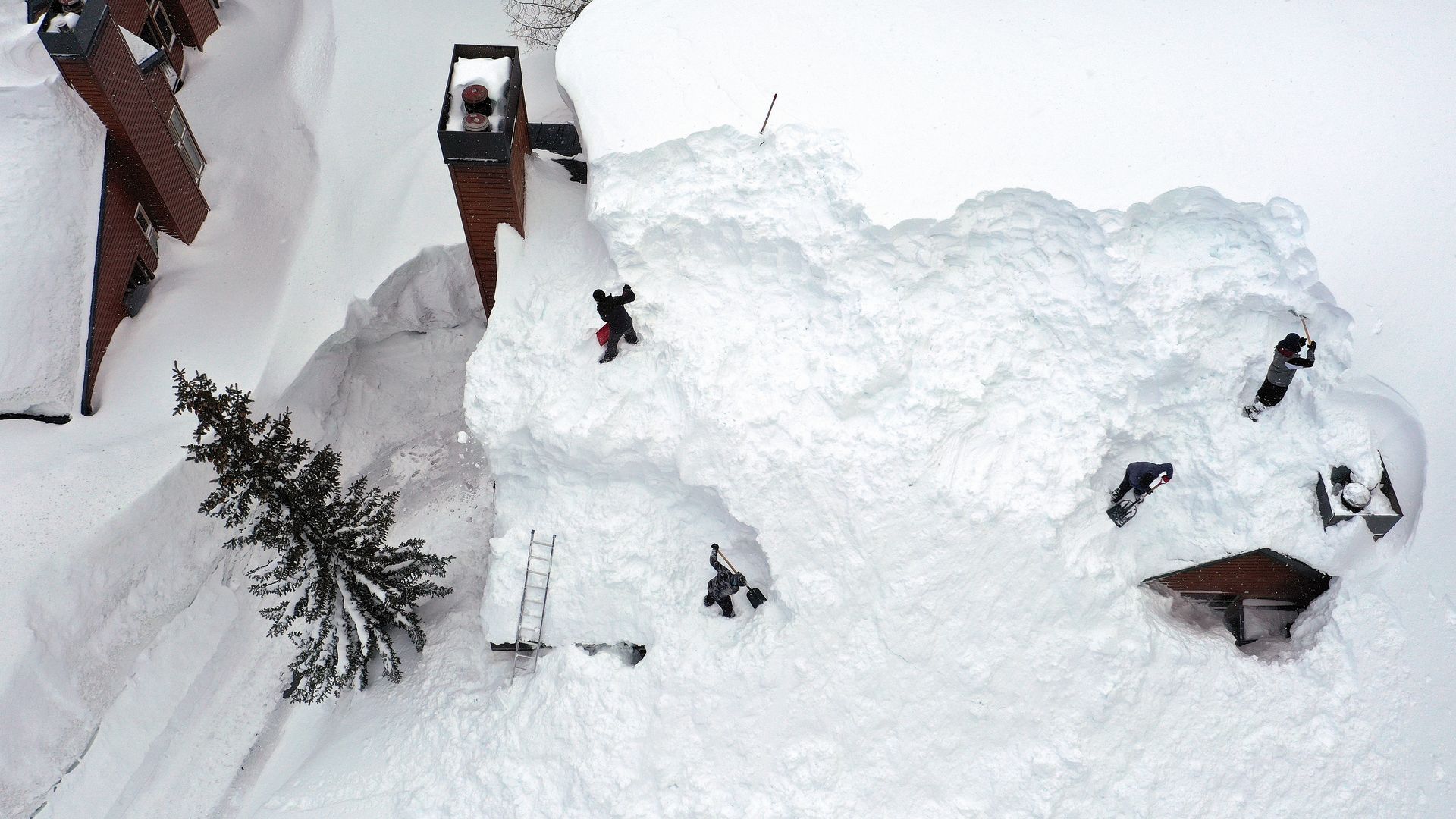Stacks of snow obscure the roof of a home; a chimney, part of a dormer and a small figure of a person clearing the snow are visible.
