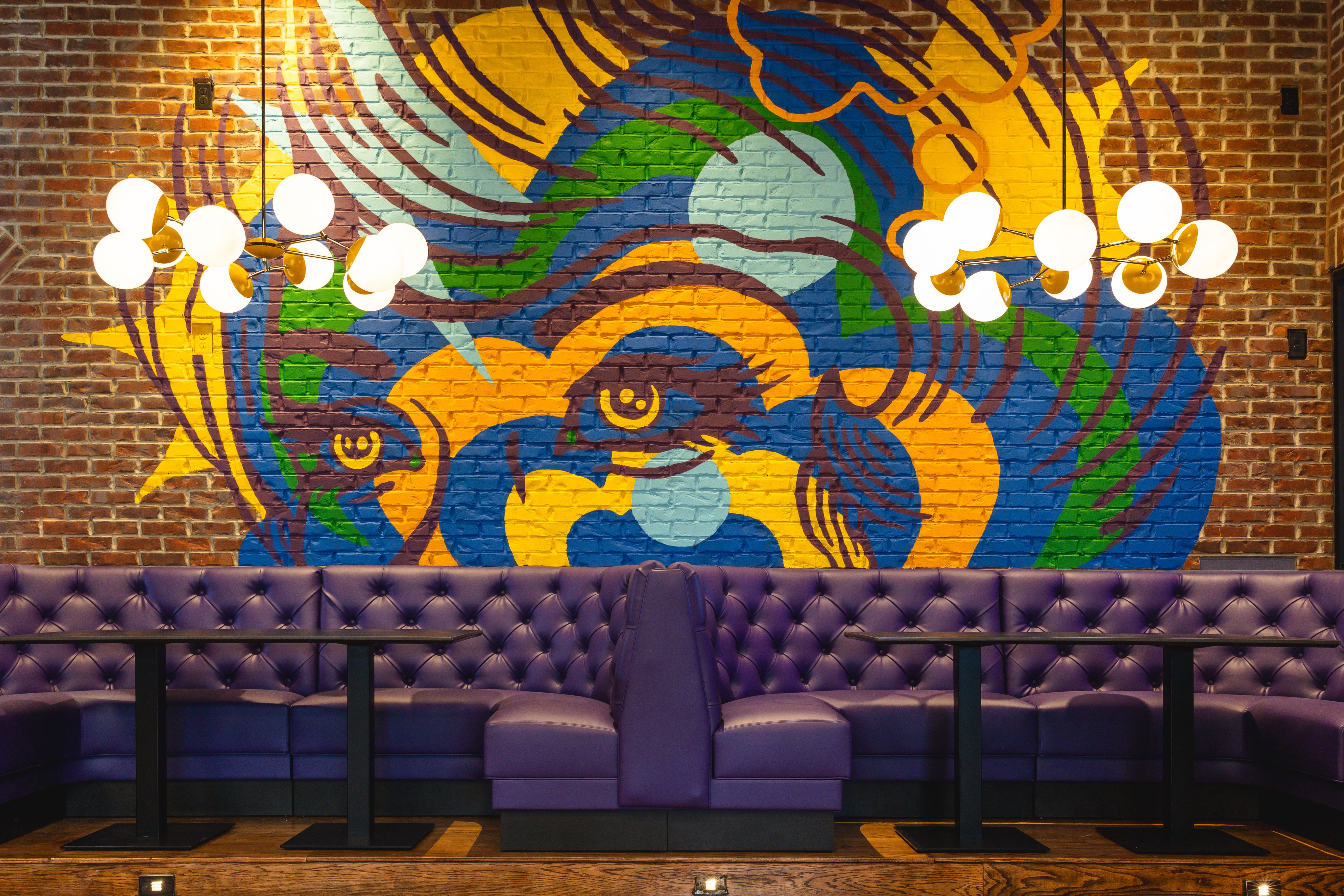 Interior of a lounge with purple tufted seating, black tables, and two modern pendant lights in front of a colorful mural of stylized eyes and abstract shapes on a brick wall.