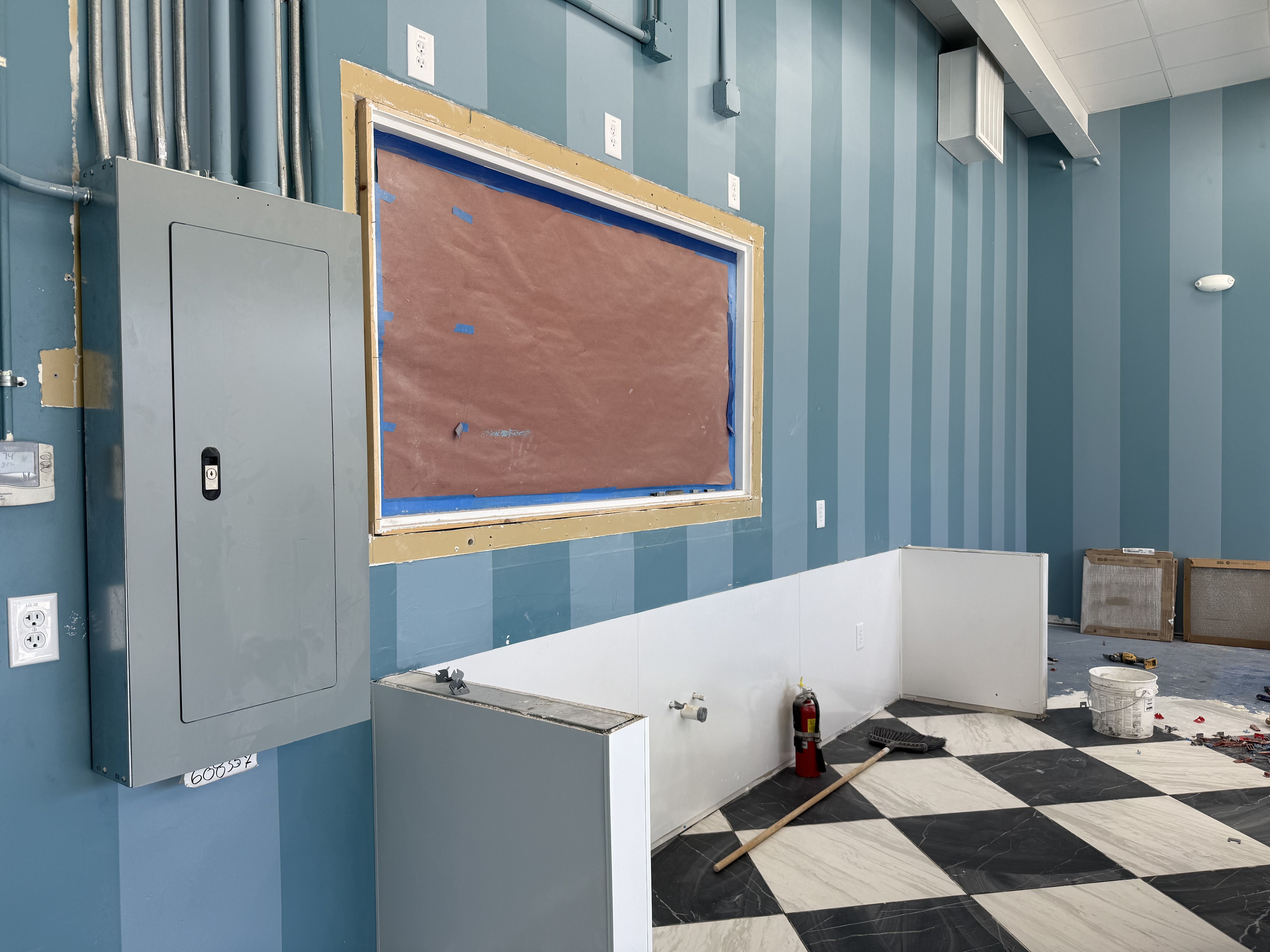 Room with blue vertical striped walls, a covered window, grey electrical panel, white paneling, black and white checkered floor, fire extinguisher, broom, and a bucket on the floor.