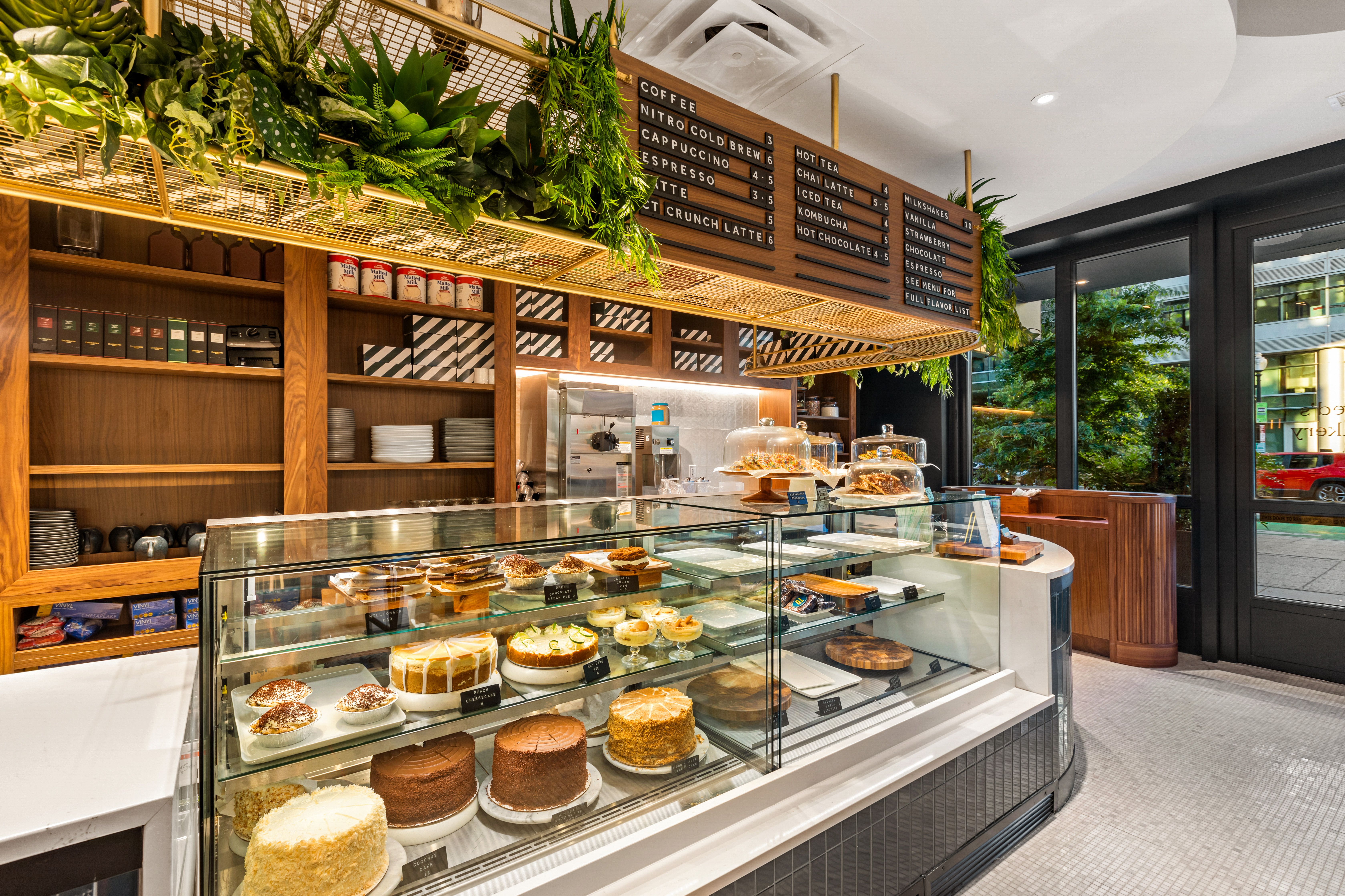 Modern cafe with a glass pastry display, cakes and pies, wooden shelves, and a wall menu. Green plants hang overhead; warm lighting and large windows reveal the street outside.