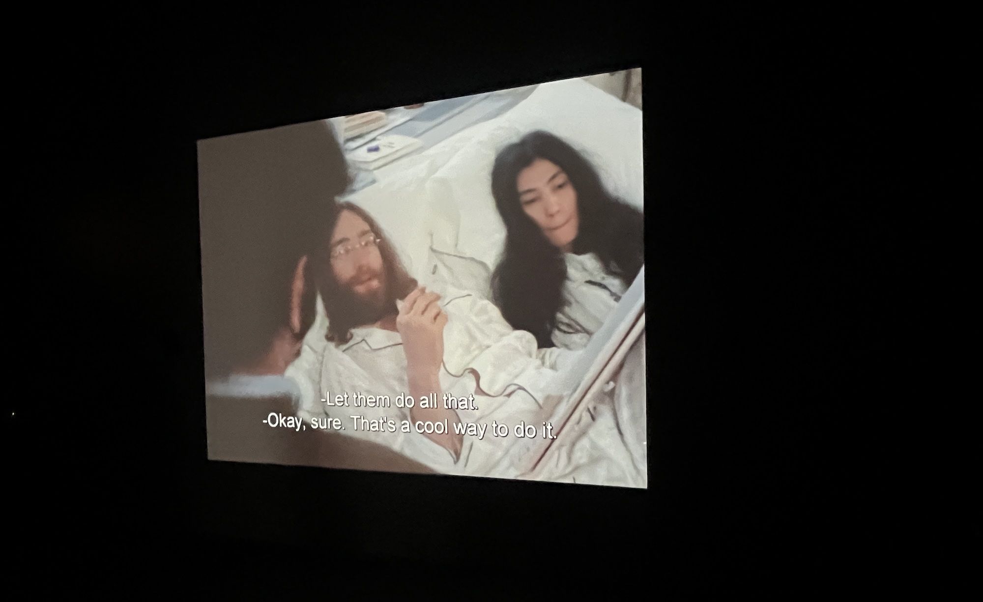 Scene from a film showing John Lennon and Yoko Ono lying on a bed in white pajamas, with subtitles that read, "Let them do all that."