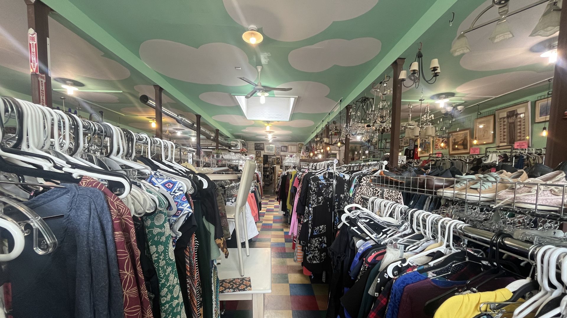 Interior of a colorful thrift store with clothing racks filled with various clothes, shoes on shelves, a green ceiling with white cloud shapes, and multicolored tiled floor.