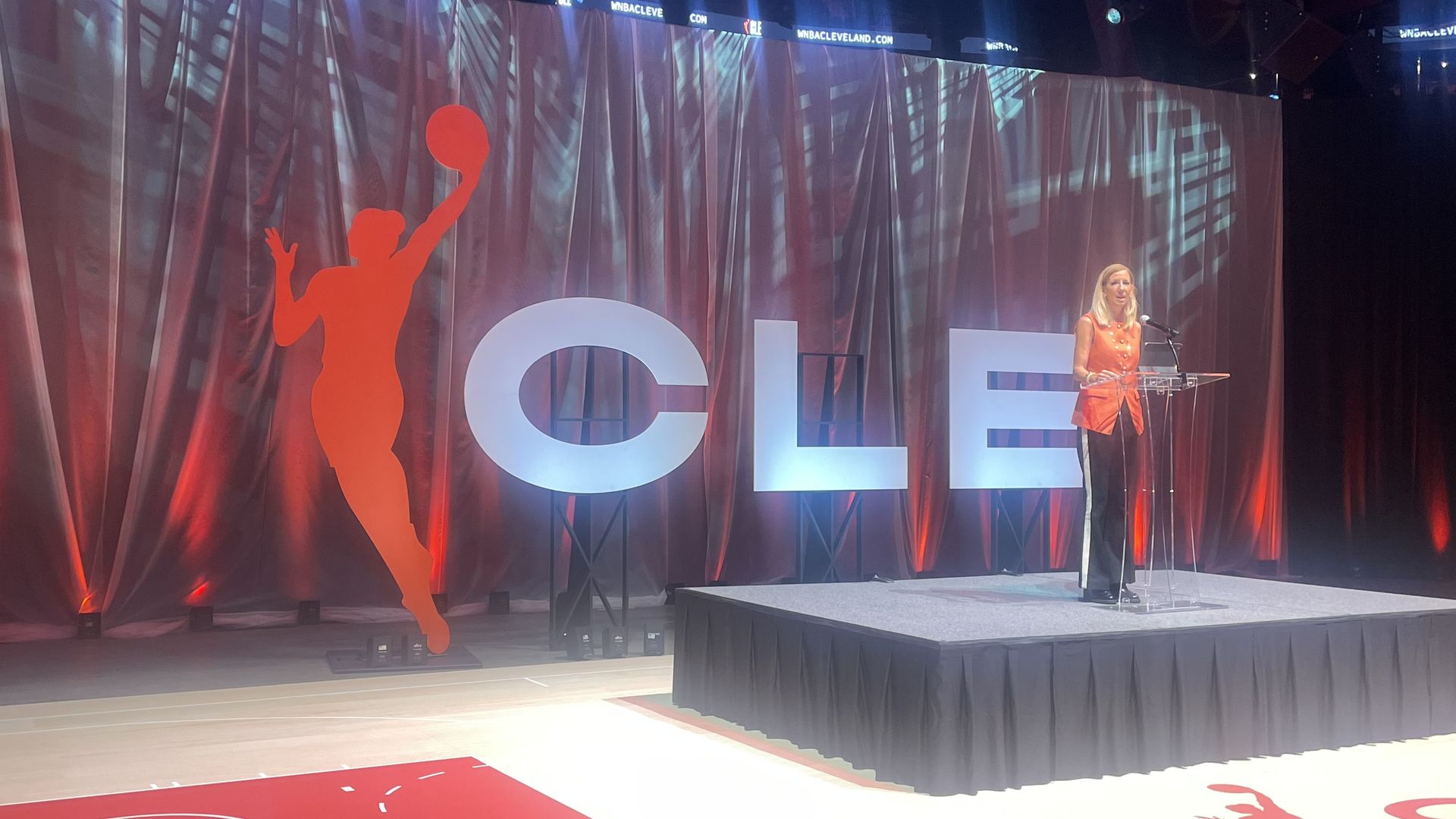 Woman in orange top speaking at a clear podium on stage with large letters "CLE" and a red silhouette of a basketball player on a dark, curtain backdrop lit with red and white lights.