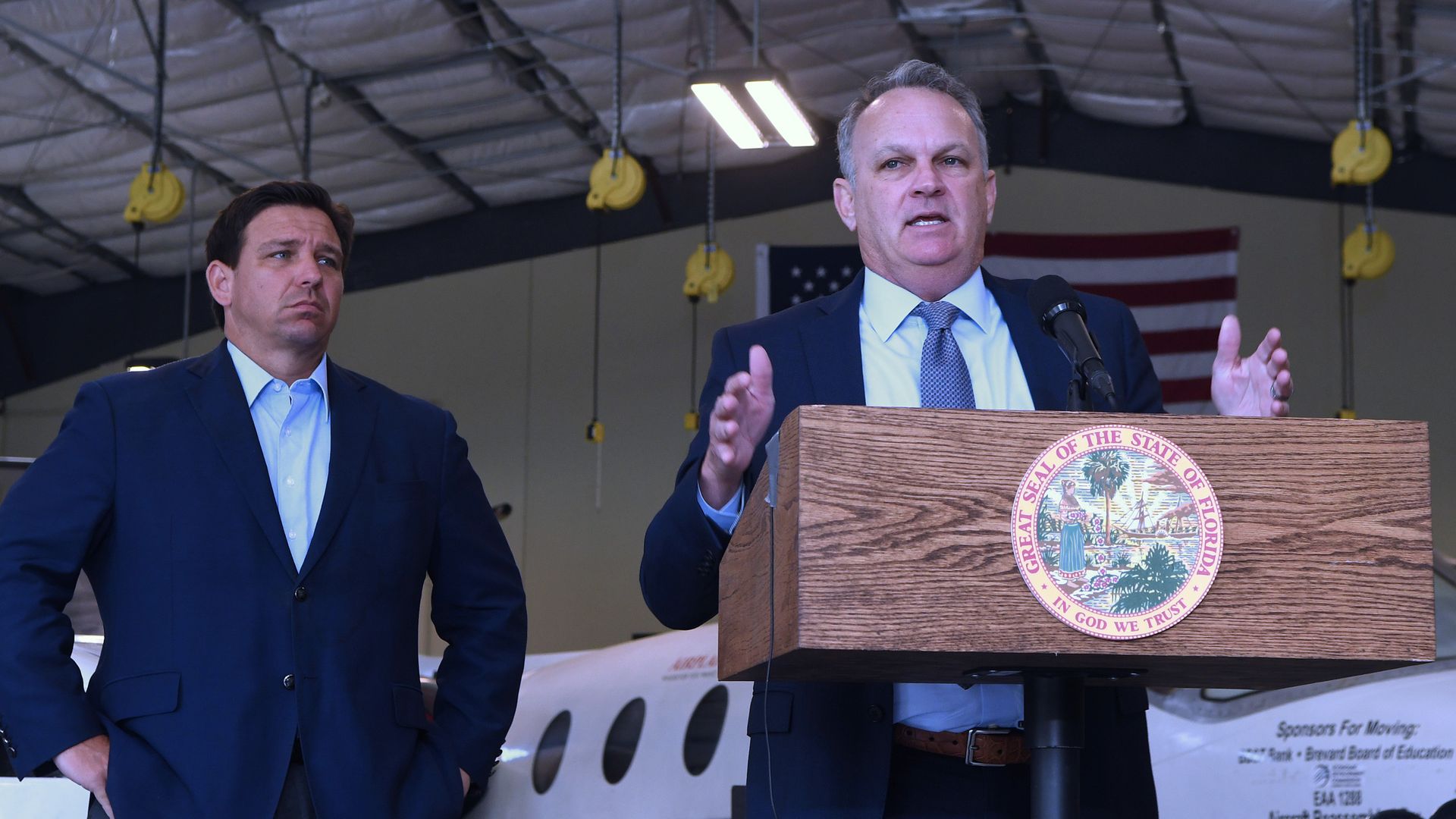 Two men in navy suits, one in front of a lectern emblazoned with the seal of Florida, speak in an airplane hangar.