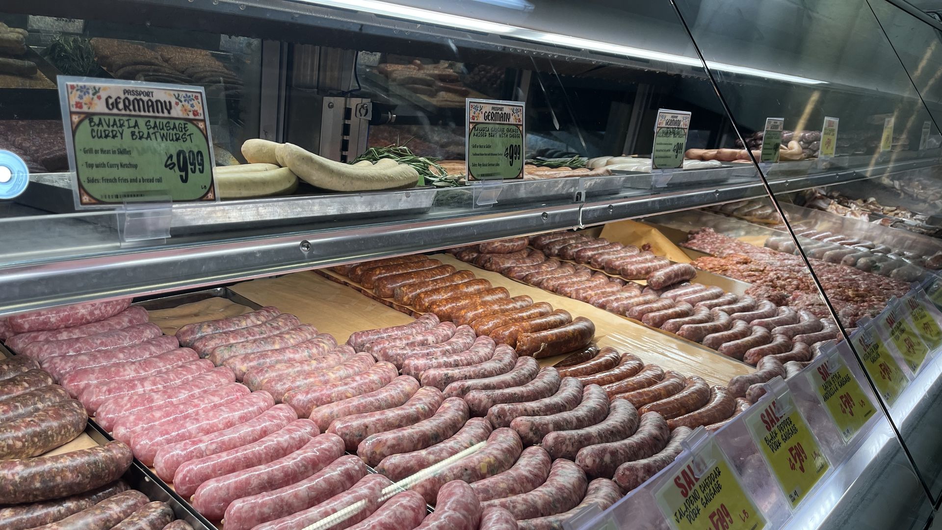 Display case filled with rows of assorted sausages including Bavaria sausage and curry bratwurst, with colorful price signs indicating sales and prices per pound.