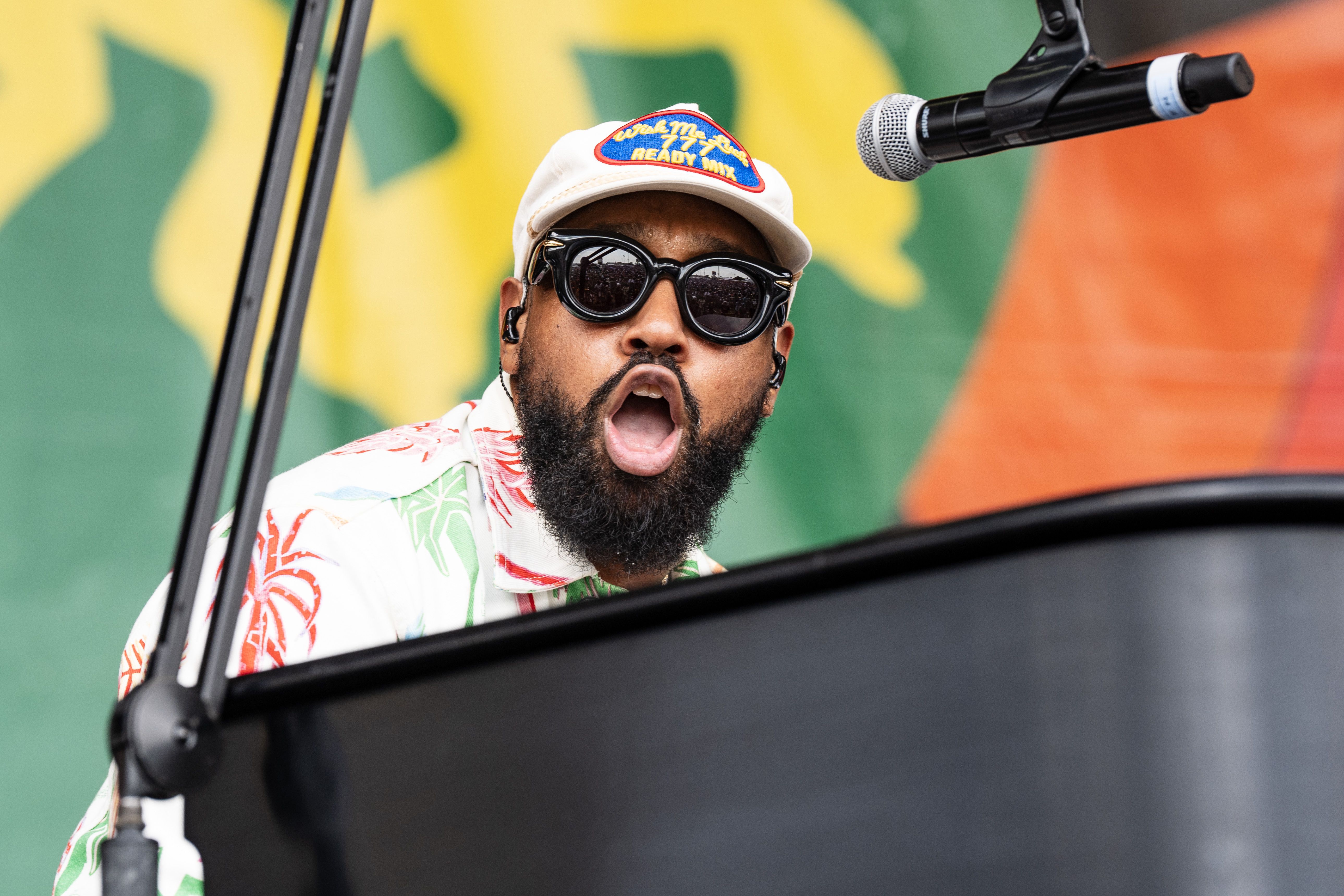 A close-up photo of PJ Morton sitting at a black piano, looking at the camera as he sings. A microphone hovers near his head.