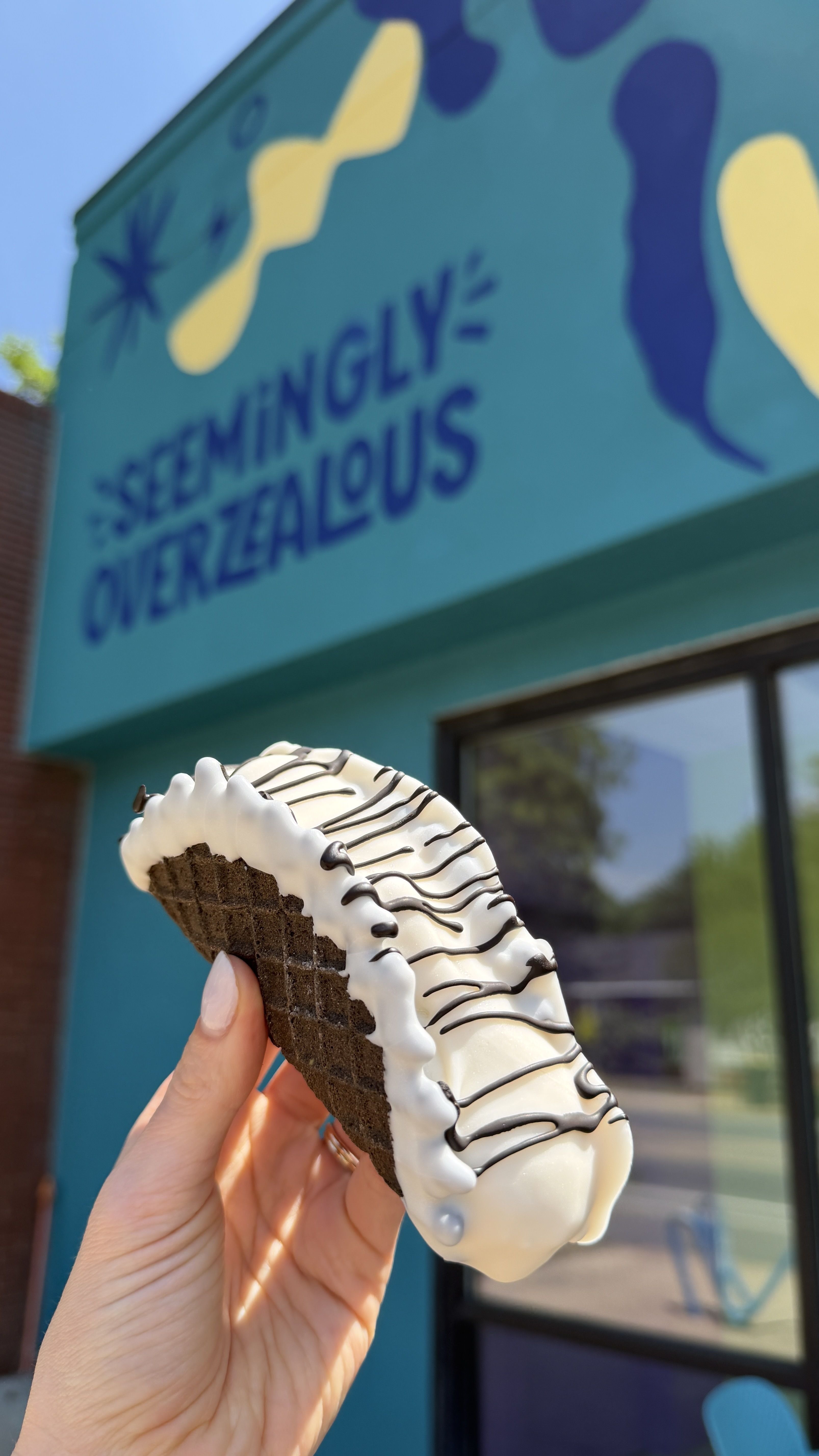 Close-up of a hand holding a white frosted chocolate ice cream cone with dark drizzle, in front of a teal storefront sign with yellow shapes and the text "SEEMINGLY OVERZEALOUS".