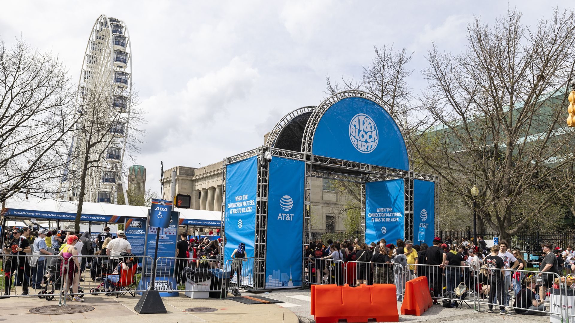 Crowd gathers around a blue AT&T "Block Party" stage on a city street, fenced by orange barriers. Leafless trees line the scene as a distant Ferris wheel looms against a cloudy sky.