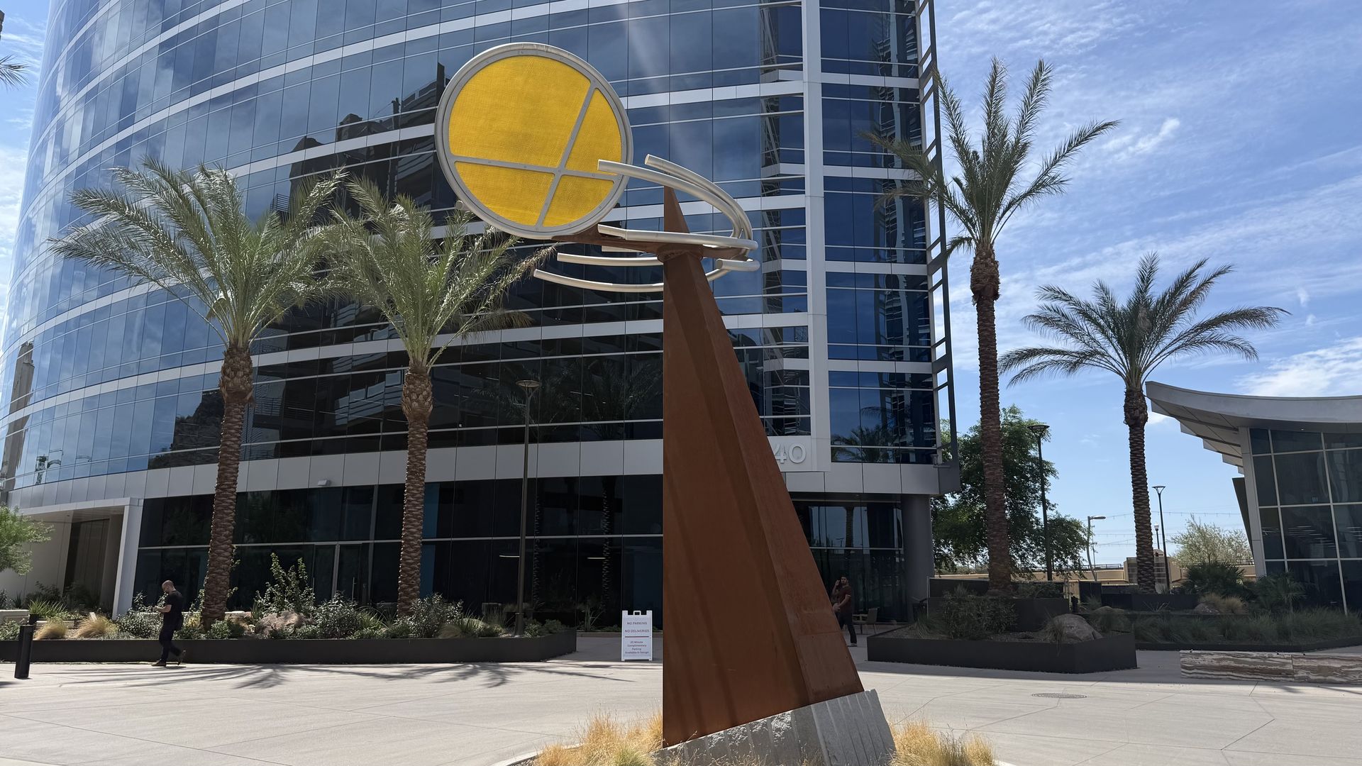 A rust-colored, elongated triangle, pointed up and tilted slightly to the side, holds a large yellow disk with silver crossbars and orbiting rings. A modern curved-glass office building with palm trees stands in the background. 