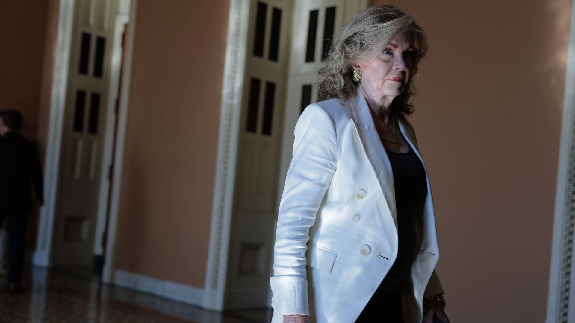 Older woman with light hair wearing a white blazer and black dress walking indoors near peach walls and ornate door frames, holding a phone in her hand.