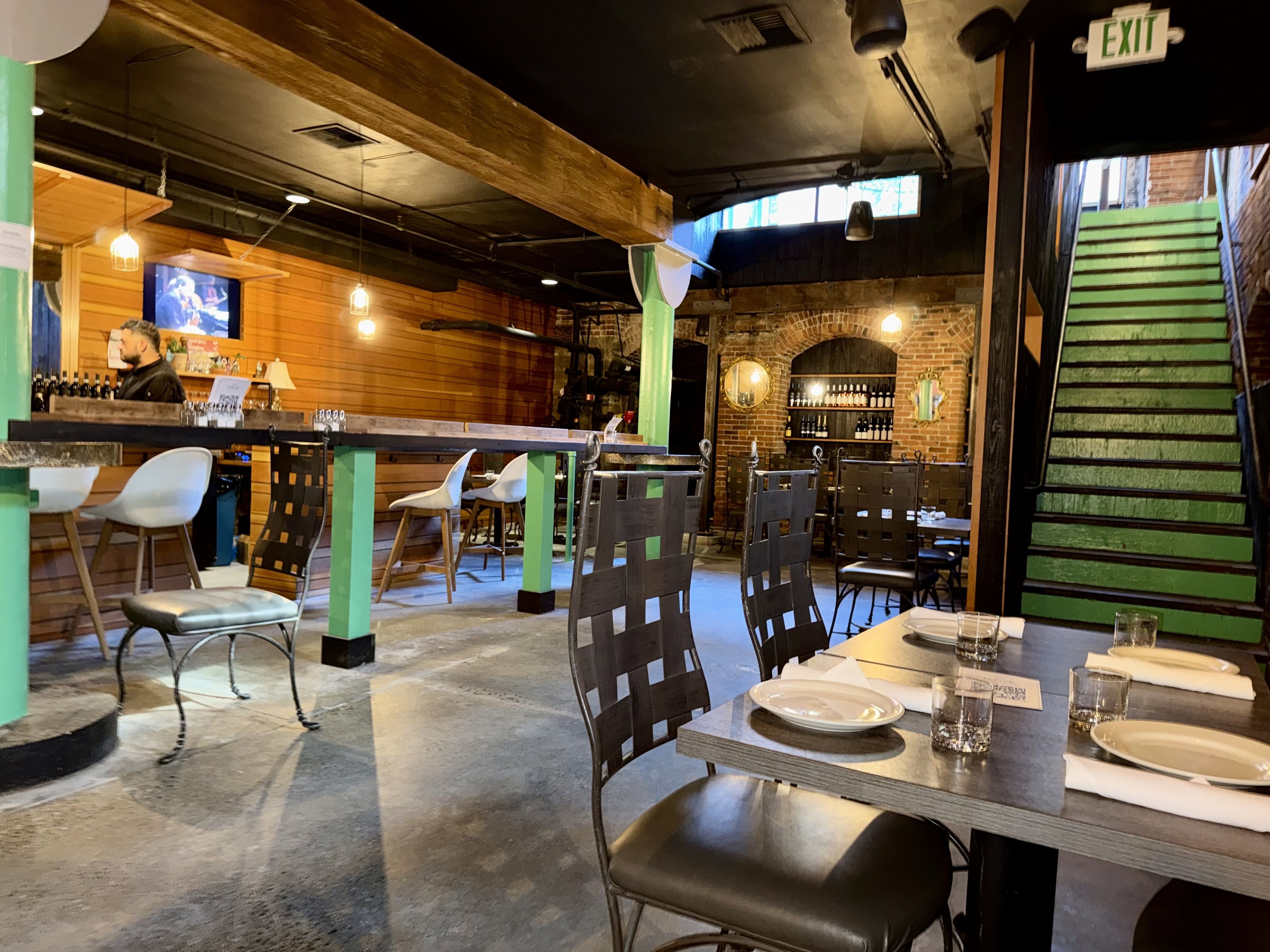 Rustic industrial restaurant interior with wooden walls and green supports. A bartender at a long bar, warm hanging lights, brick arches, bottled shelves, and a green stairway by an EXIT sign.
