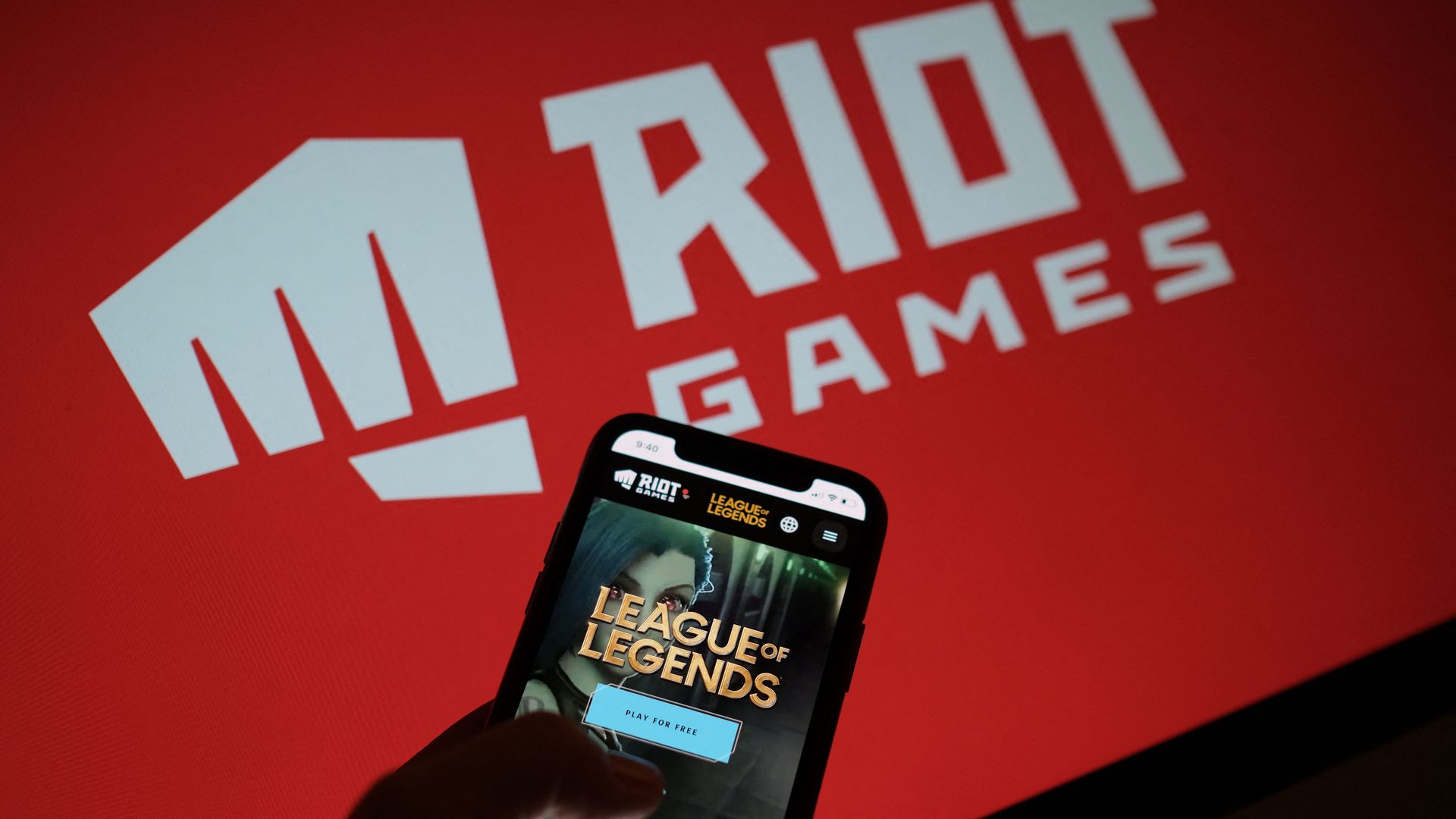 Photo of a phone showing the logo of League of Legends, held in front of a logo for Riot Games
