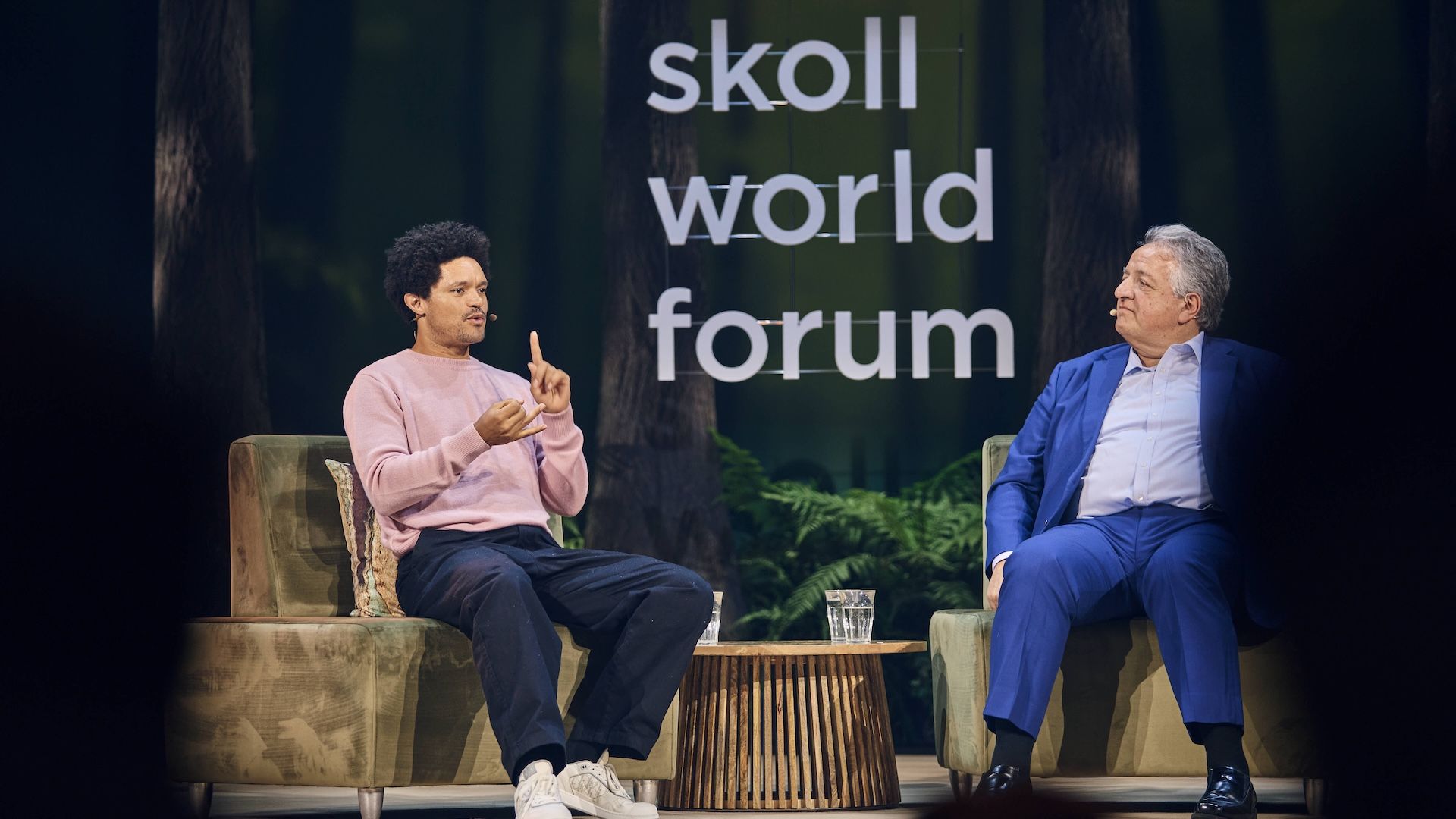 Two men sit on a stage in front of a forest backdrop at the Skoll World Forum; a large sign reads "Skoll World Forum." One wears a pink sweater and gestures, the other in a blue suit listens.